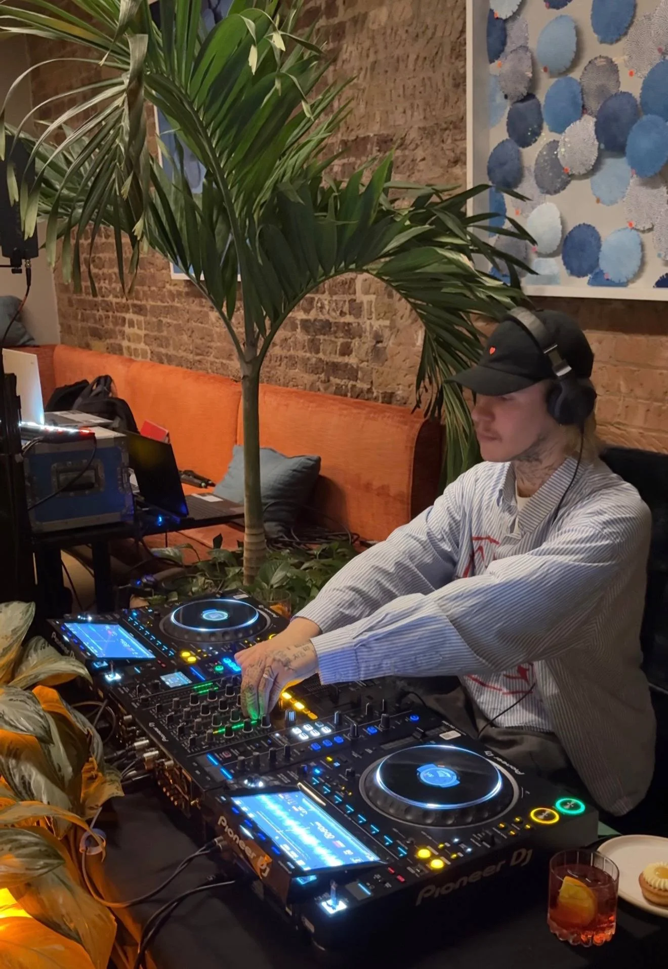 A DJ wearing a striped shirt and a black cap is performing at a DJ booth with Pioneer equipment, headphones on, in a room with exposed brick walls, a large indoor plant, and abstract art on the wall. A glass of orange drink is on the table.