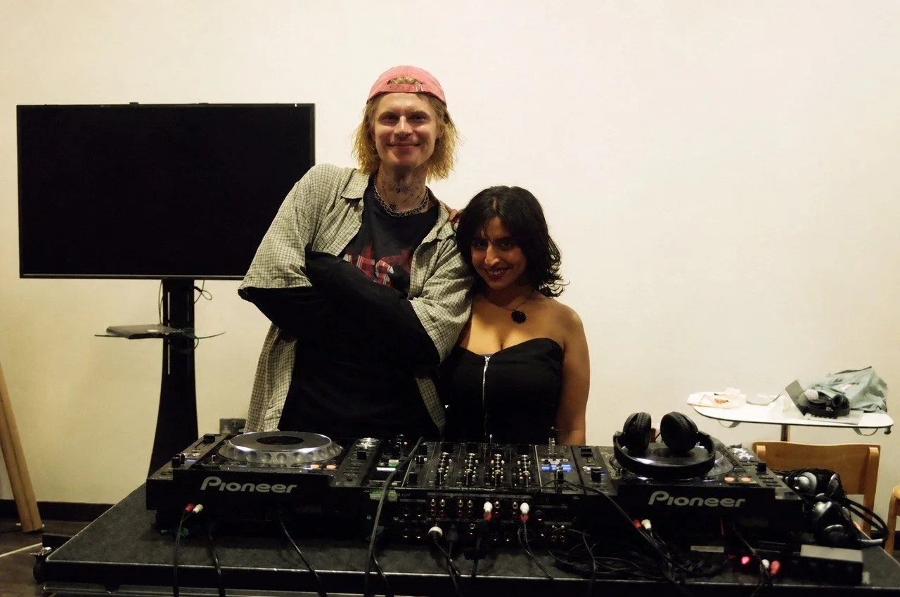 A man and woman are standing behind DJ equipment, smiling at the camera in a room with a plain white wall. The man has long blond hair, wears a pink cap, a checkered shirt, and a black t-shirt, while the woman has shoulder-length dark hair, wears a black strapless top, and a necklace. There is a flat-screen TV and a small table with headphones and a bag on it in the background.