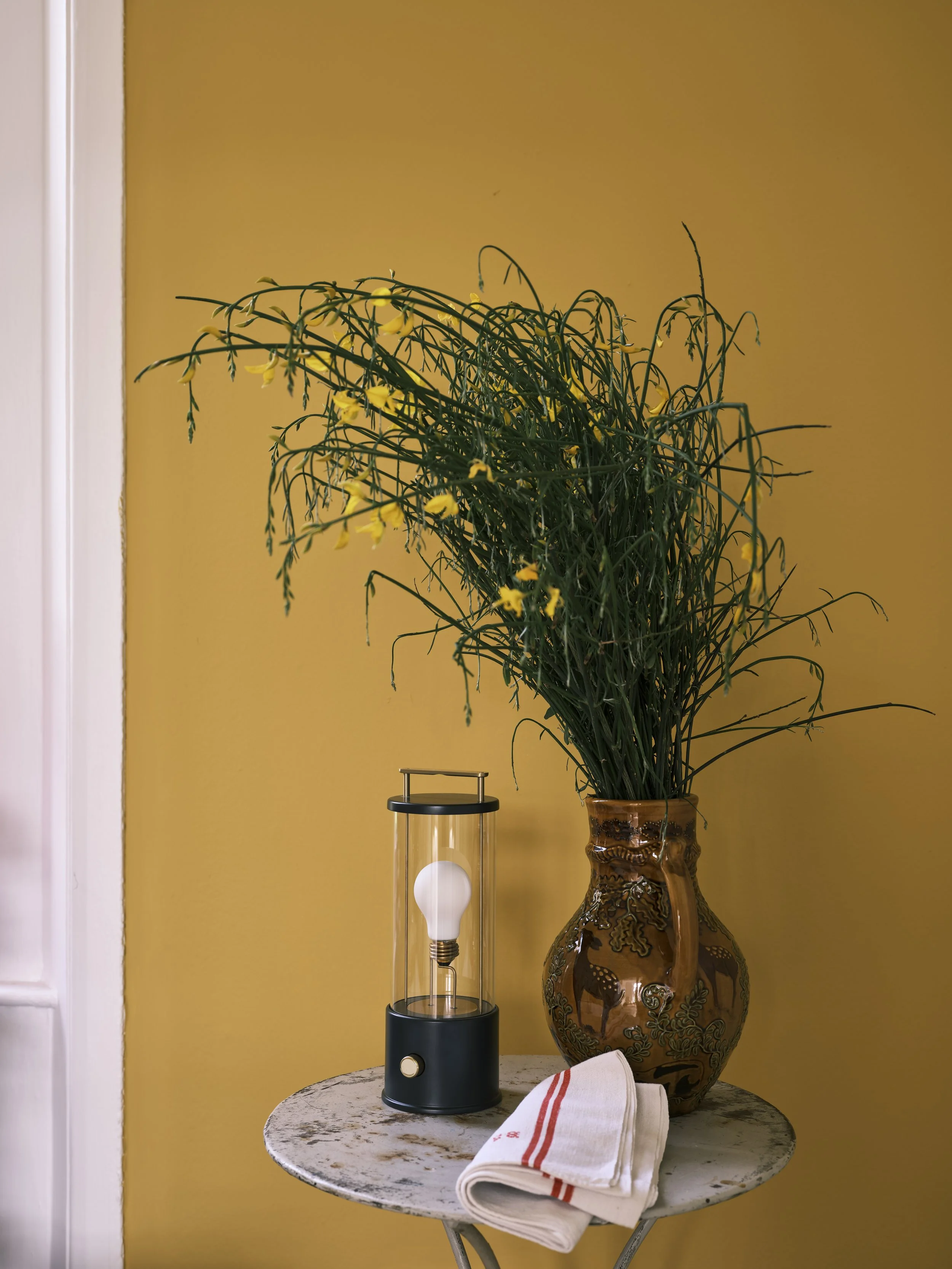 Tala portable cordless lamp on a small table beside a potted plant, set against an ochre wall.