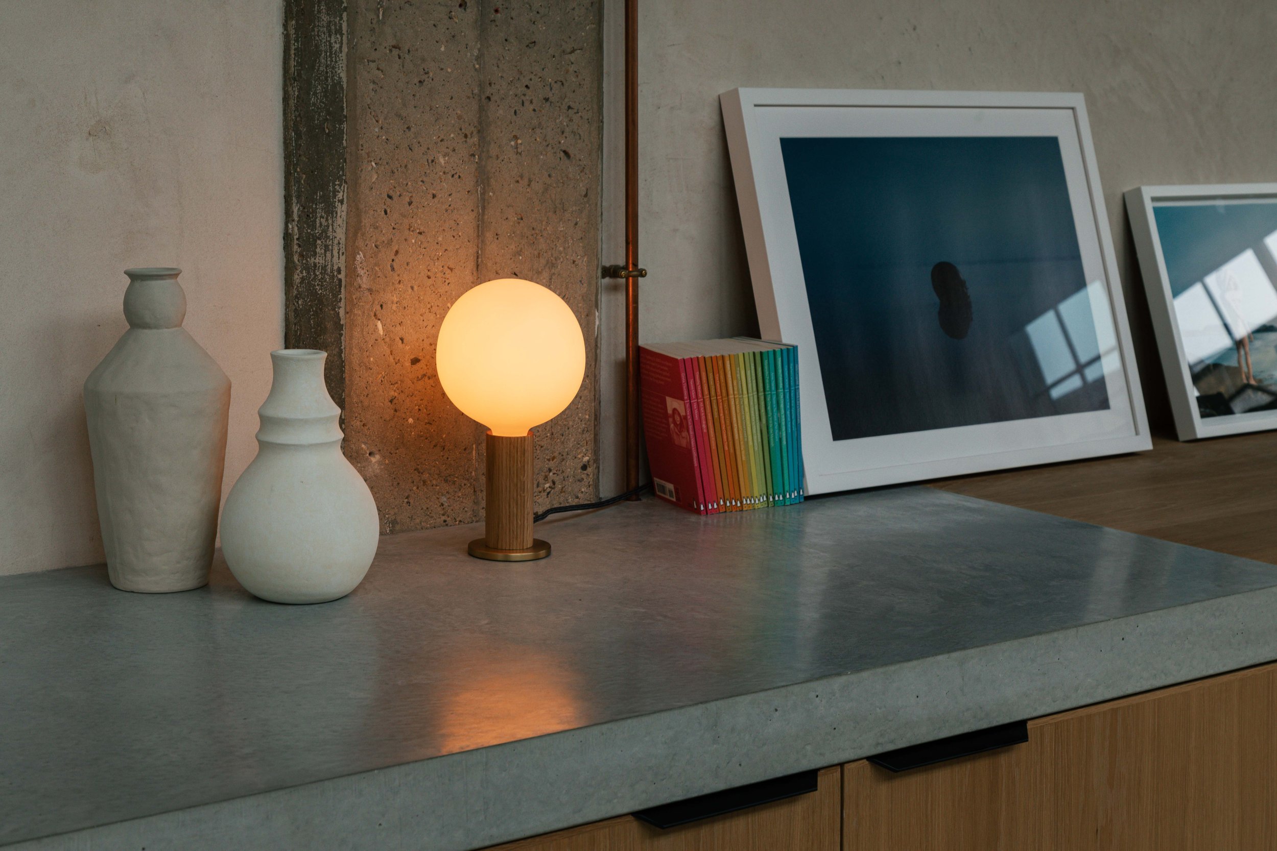 Tala lamp on a countertop alongside a decorative vase, books, and artwork against an exposed concrete wall.