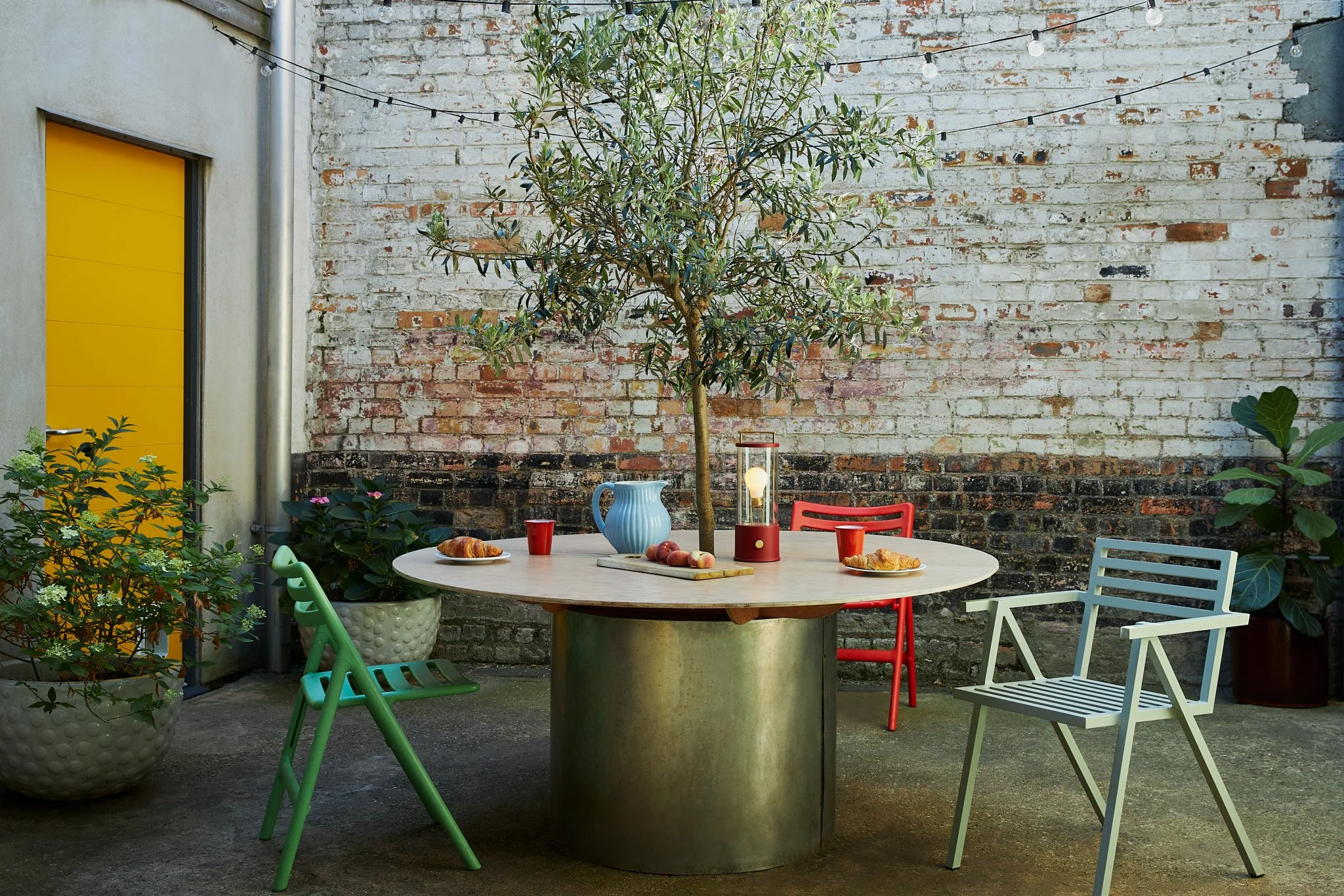 Rustic outdoor dining table featuring colourful chairs, a central olive tree, and a red Tala cordless lamp.