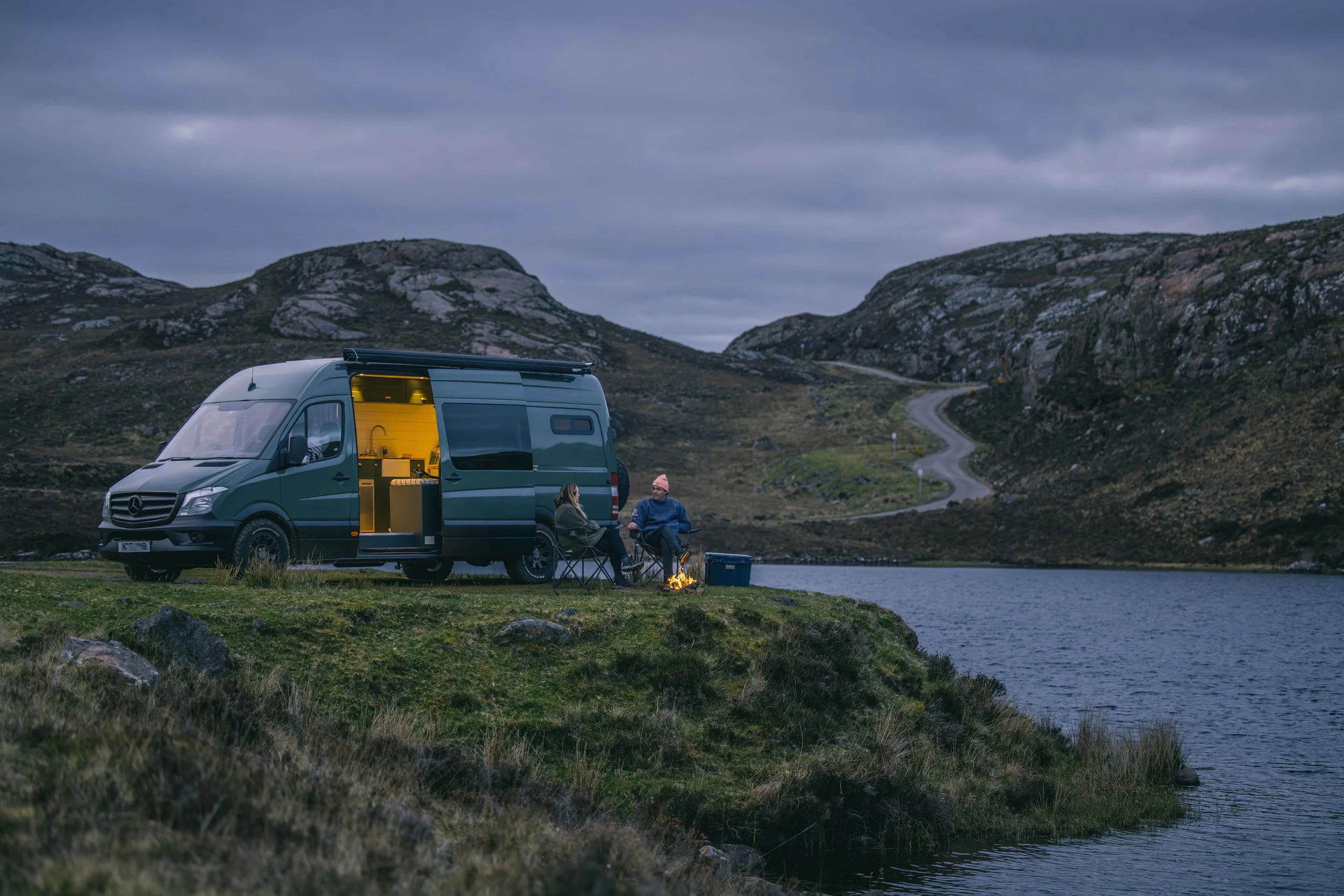 A Wingbeat Escapes camper van parked by a Scottish loch at dusk with customers sitting outside.