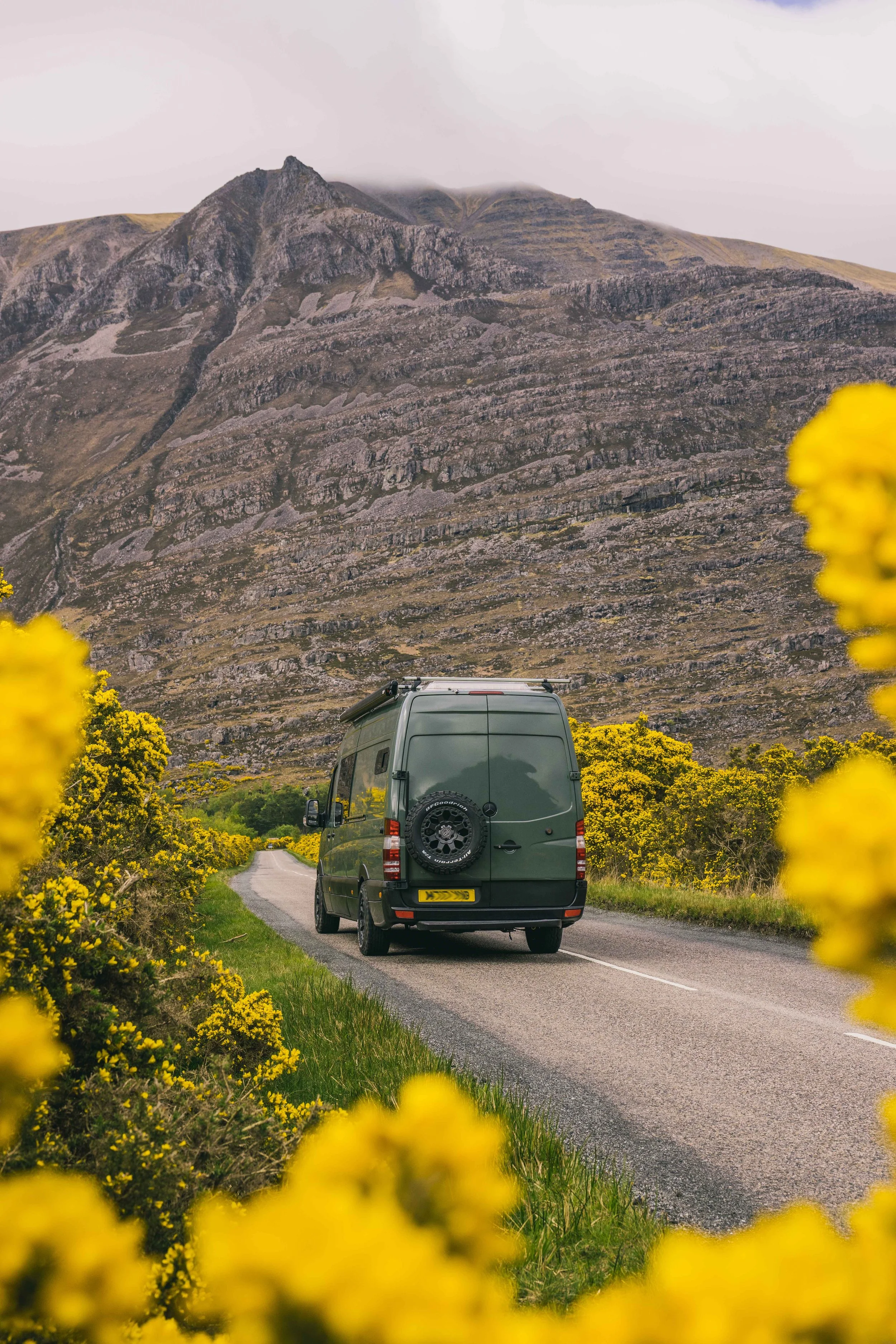 A Wingbeat Escapes camper van driving through a rugged Scottish Highlands landscape with rocky terrain.