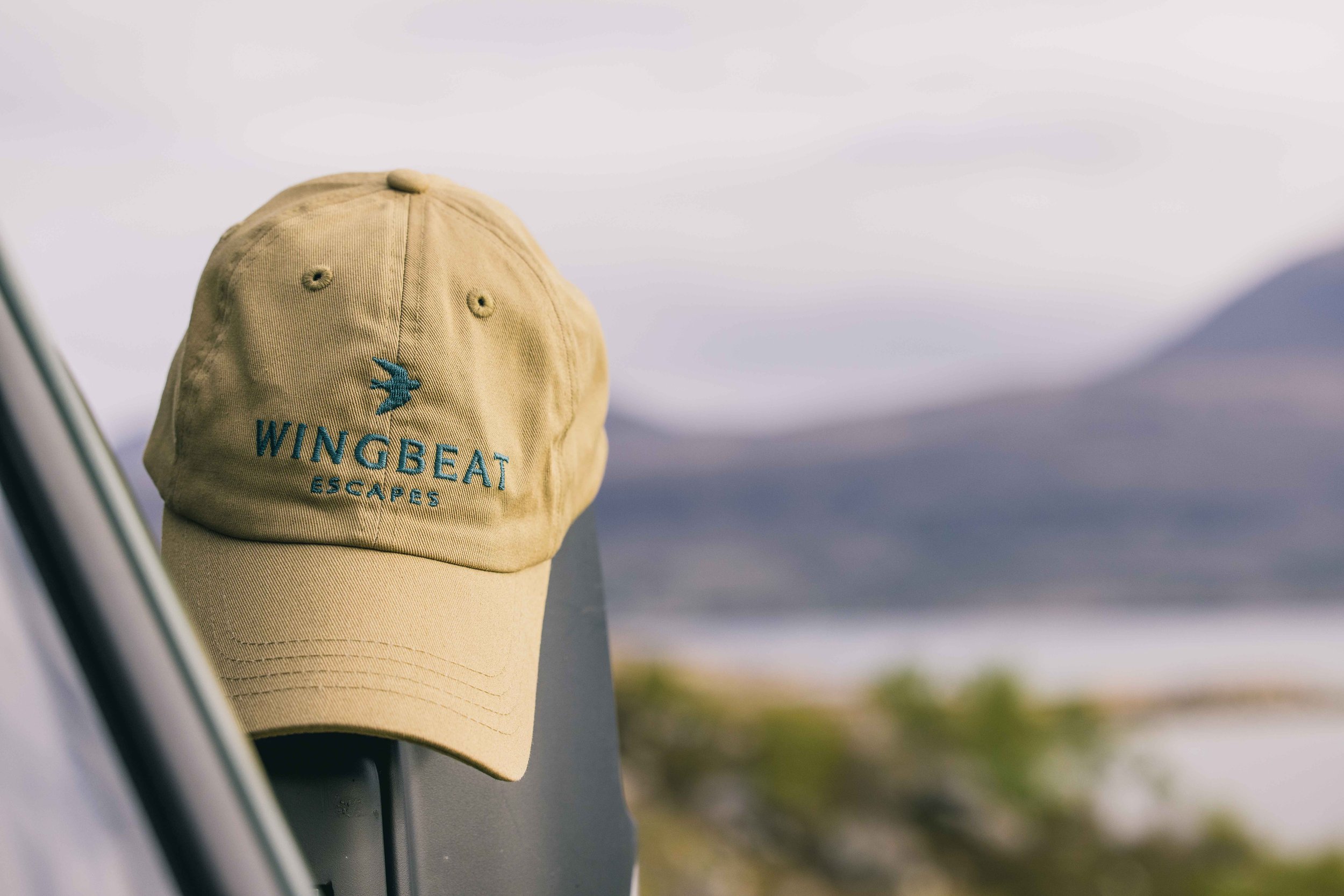 A branded Wingbeat Escapes tan cap resting elegantly on a camper van wing mirror.