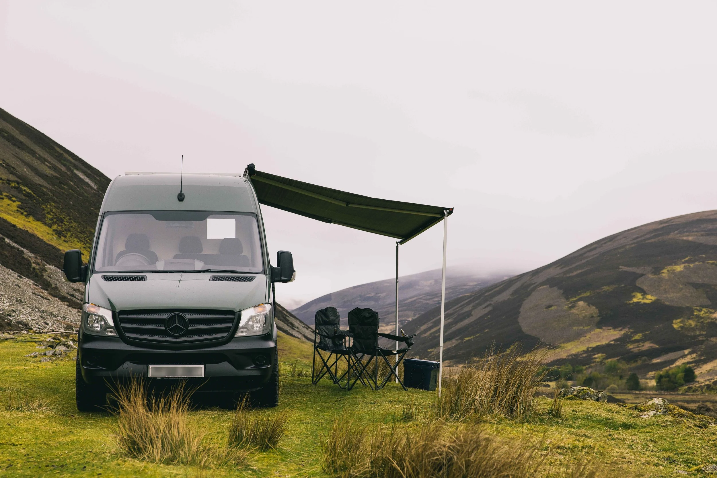 A Wingbeat Escapes camper van parked on a grassy plateau with its built-in canopy extended against misty hills.