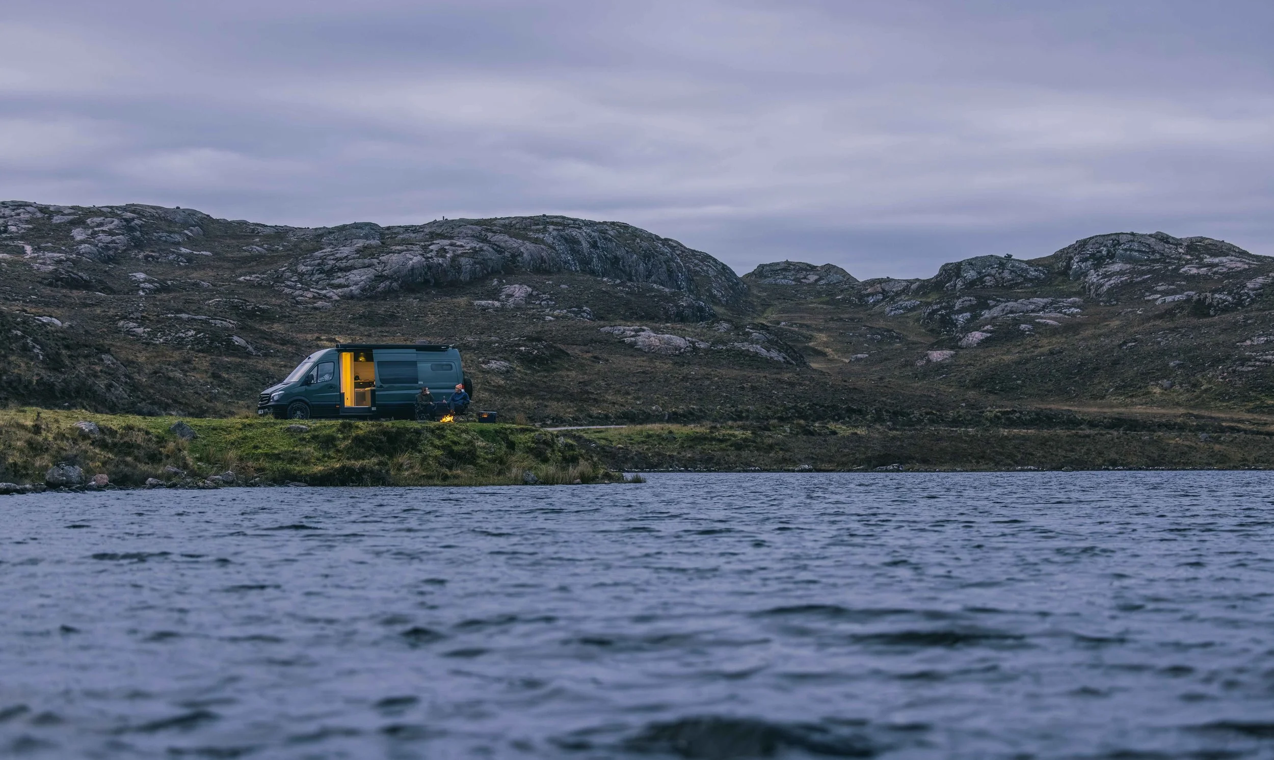 Distant view of a Wingbeat Escapes camper van parked by a Scottish loch at dusk with a warmly lit interior.