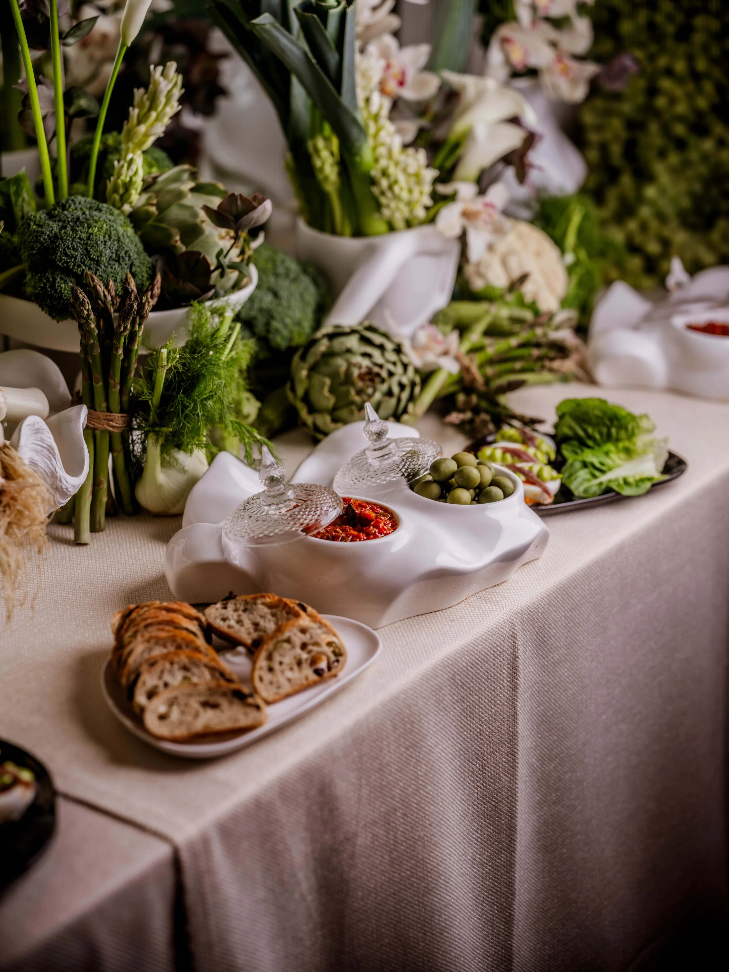 An elegant banquet table styled with artisanal nibbles, decorative green vegetables, and curated floral displays.
