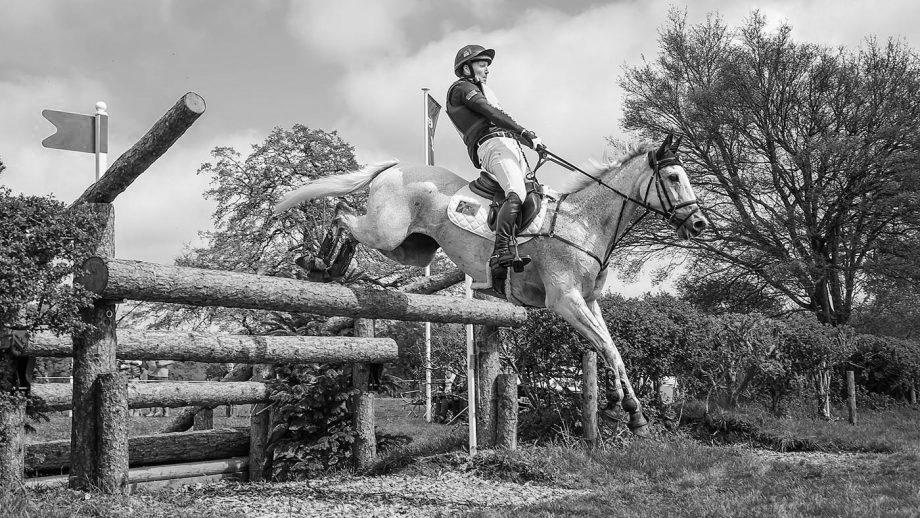 Black and white shot of a horse and rider clearing a rustic wooden log jump at an event.