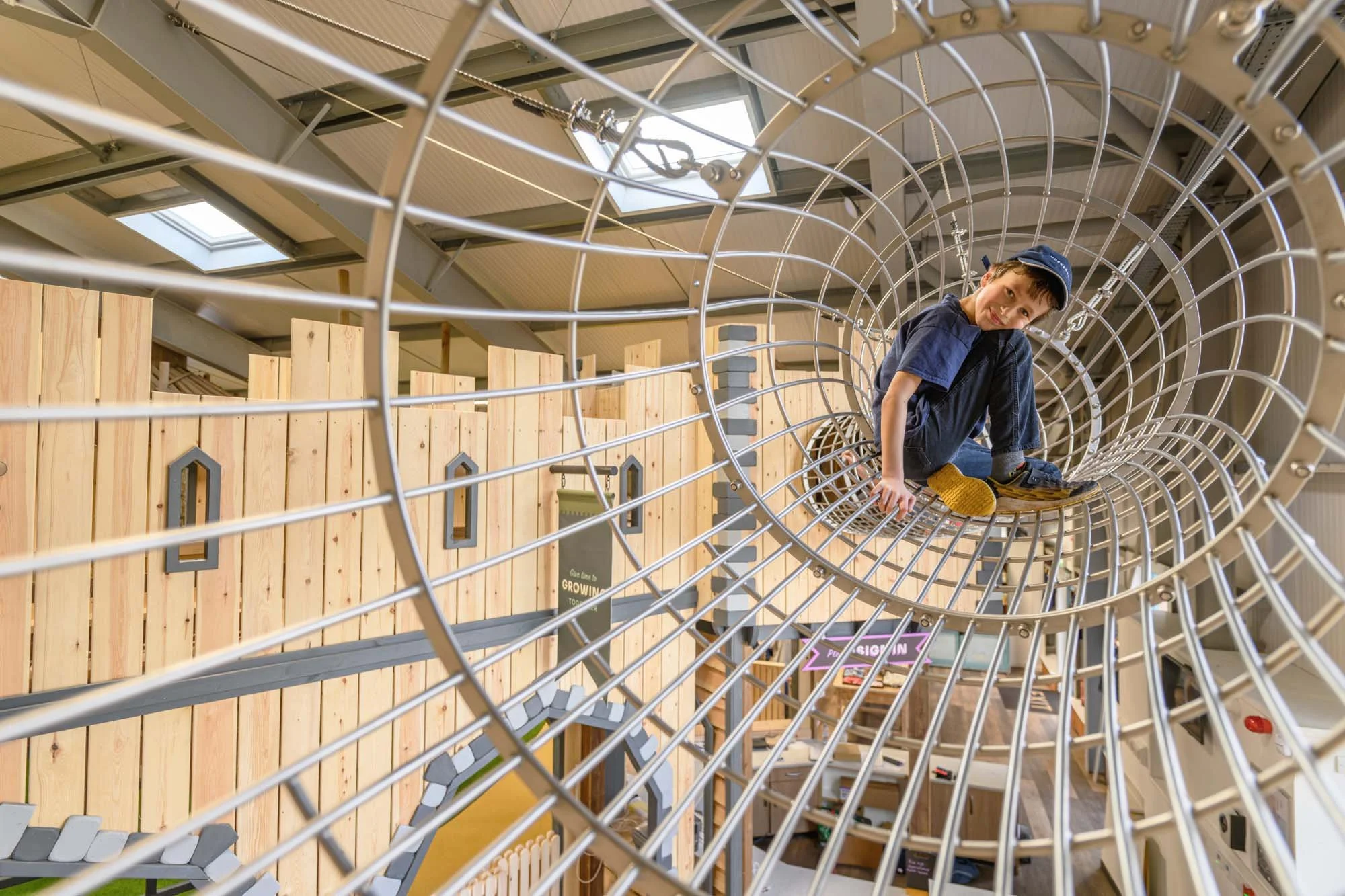 A smiling child sitting inside the elevated cylindrical wire crawl-bridge at Hockerhill Playbarn.