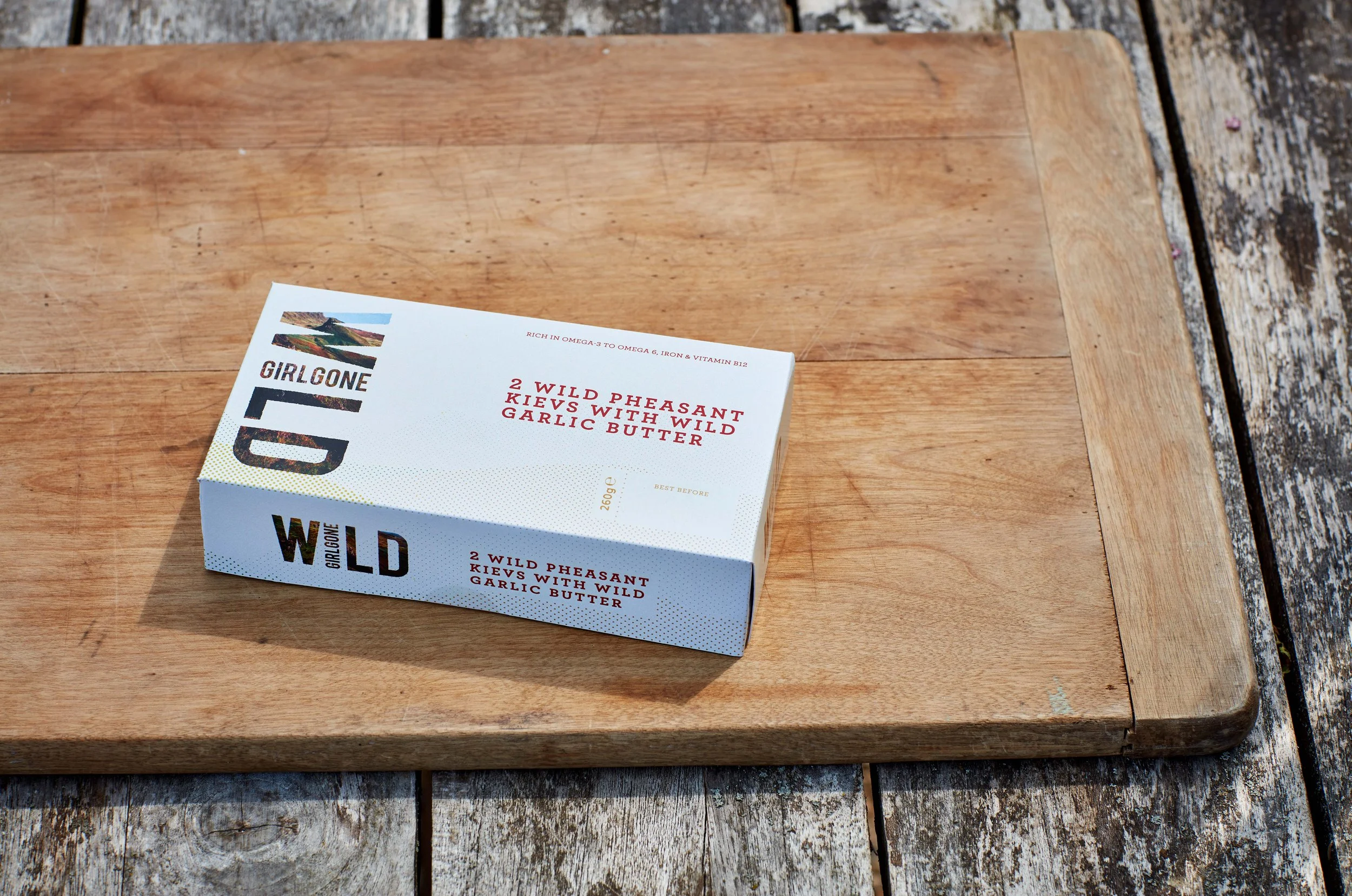 Girl Gone Wild Pheasant Kiev packaging resting on a wooden chopping board atop an outdoor dining table.