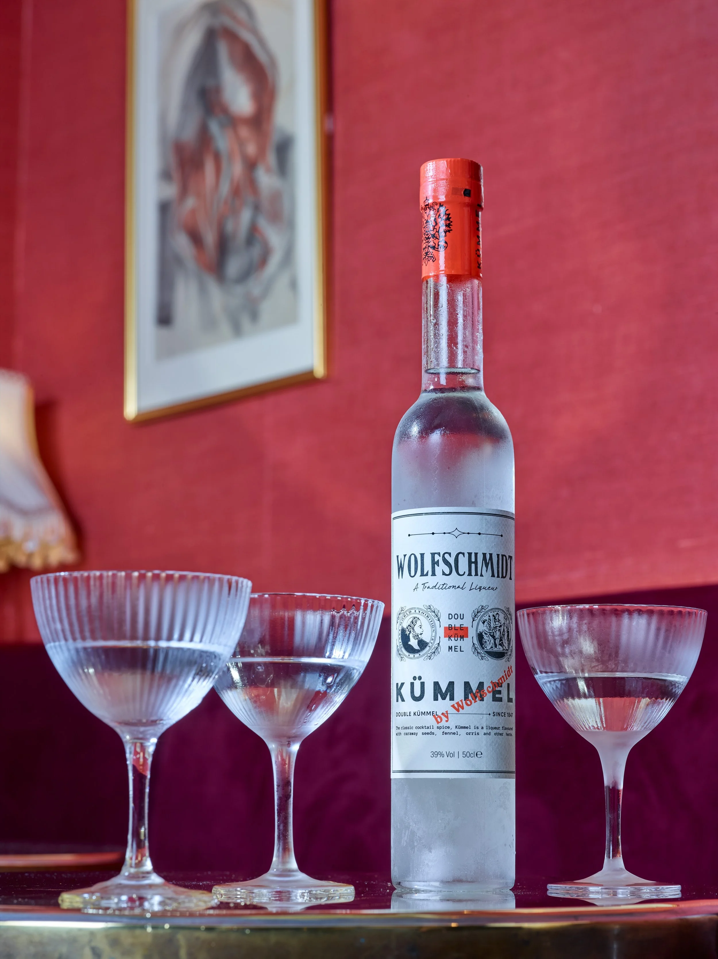 An ice-cold Wolfschmidt Kümmel bottle paired with elegant coupe glasses, set against a vibrant red backdrop.