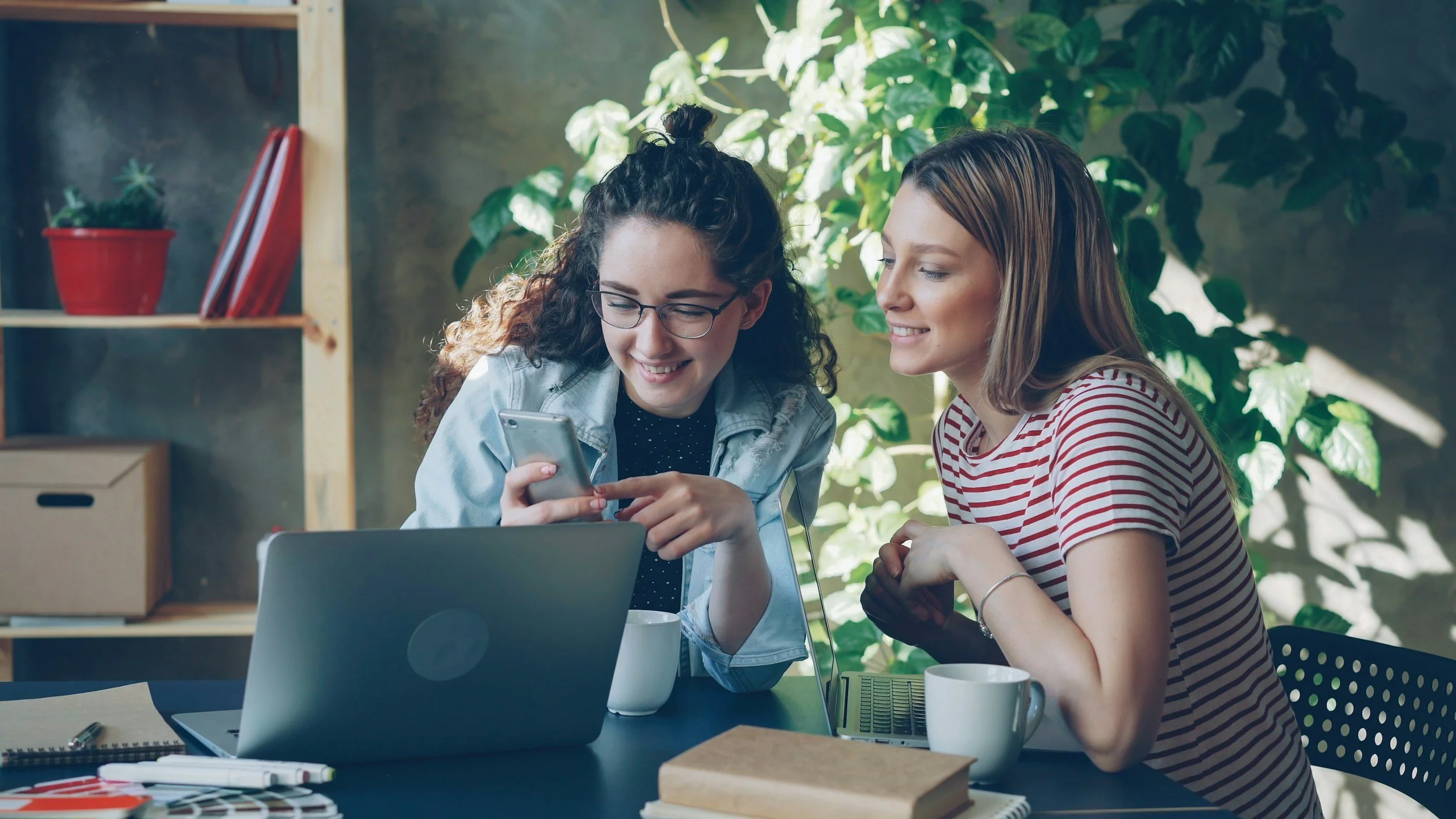 Two young women sit at a table in a bright room with greenery, looking at a smartphone together, smiling and engaging with each other.