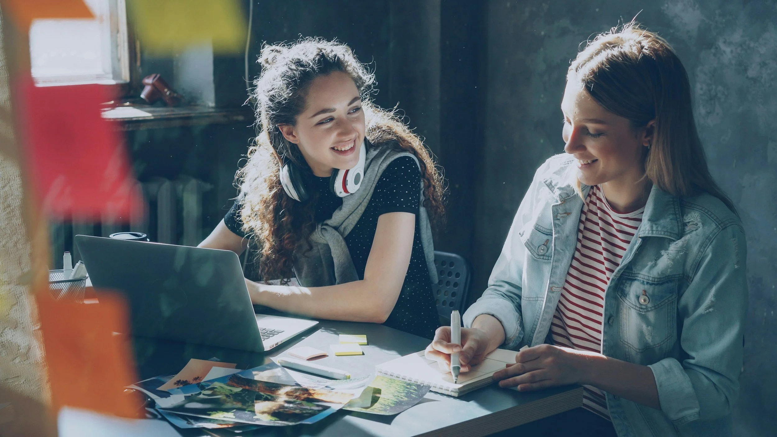 Two young women sit at a desk in a well-lit room, smiling and engaging in a discussion. One is wearing a gray jacket and striped shirt, writing in a notebook, and the other is wearing a dark top with a gray sweater tied around her shoulders and has headphones around her neck. There's a laptop and various papers on the desk.
