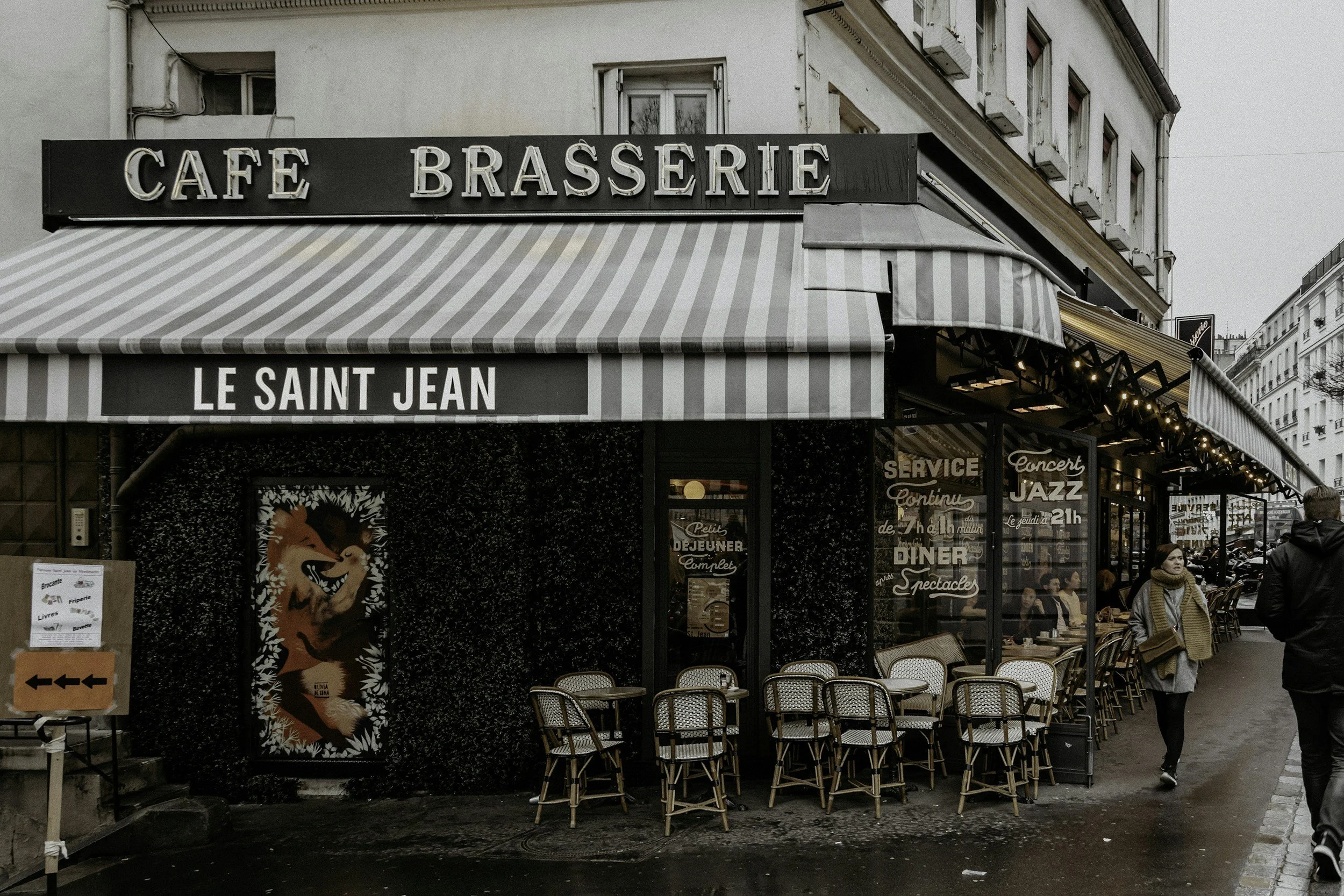 Street view of a cafe and brasserie named 'Le Saint Jean' with outdoor seating, striped awning, and several people walking by.