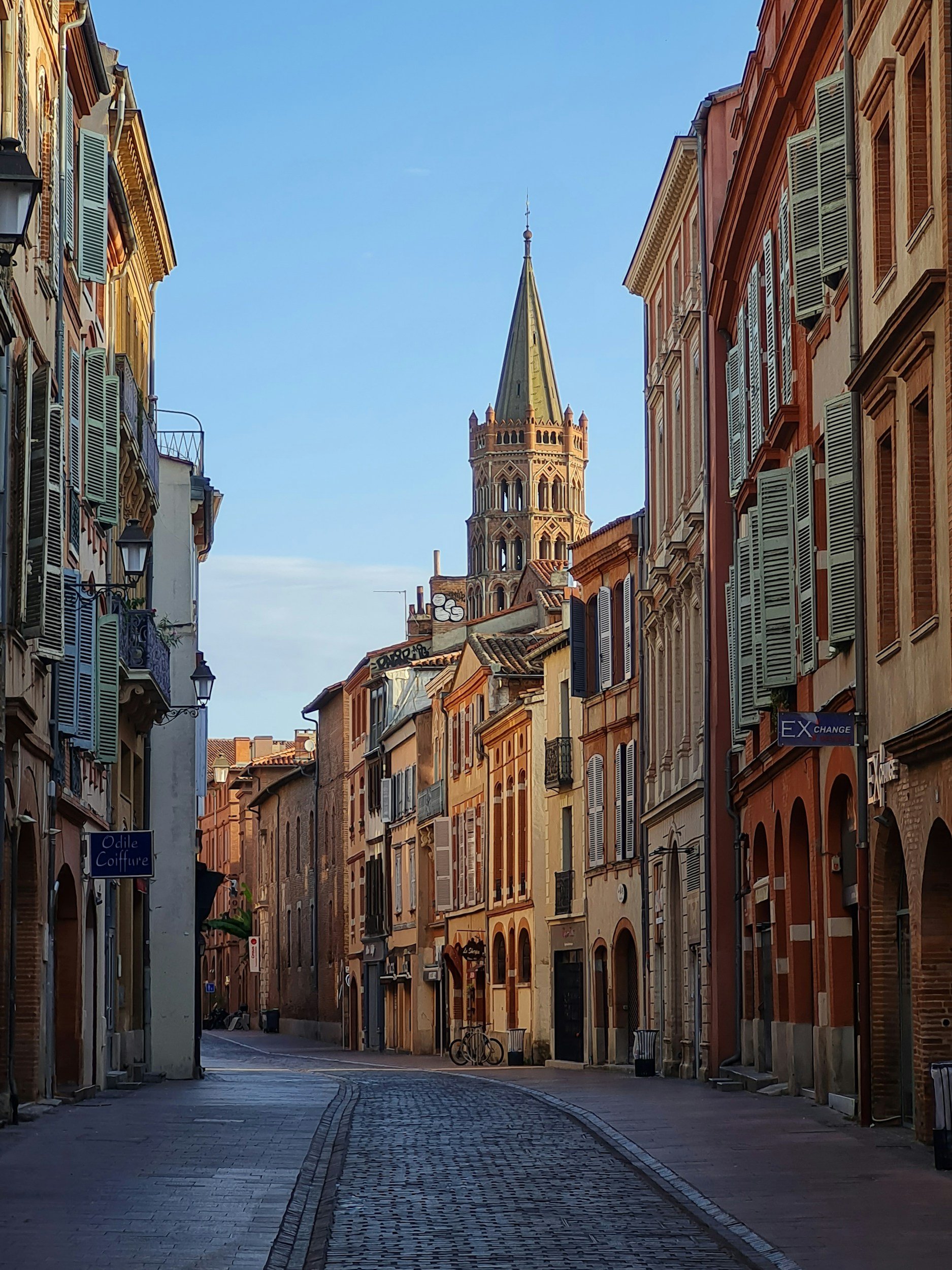 A narrow cobblestone street lined with historic European-style buildings with colorful facades, shutters, and balconies. A church tower with a spire is visible in the distance against a clear blue sky.