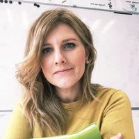 A woman with shoulder-length light brown hair, wearing a mustard-colored top, holding a green notebook, standing in a classroom.