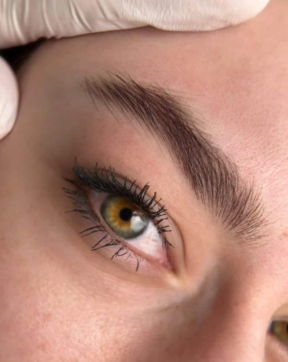 Zoom sur un œil avec un regard de côté, mettant en évidence la paupière, les cils et le sourcil, avec un iris marron clair et un fond de peau claire.