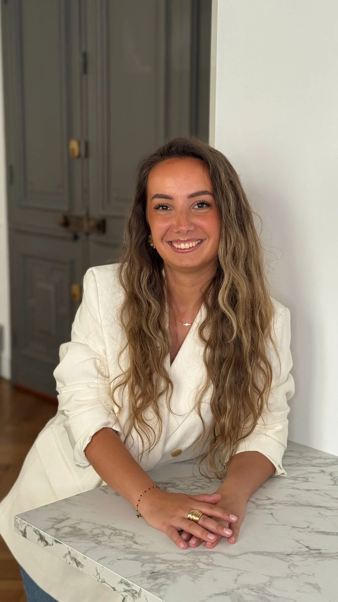 Une femme souriante avec des cheveux longs et bouclés, portant une veste blanche, posant avec ses bras croisés sur une table en marbre gris et blanc. En arrière-plan, il y a un mur blanc et des armoires grises.