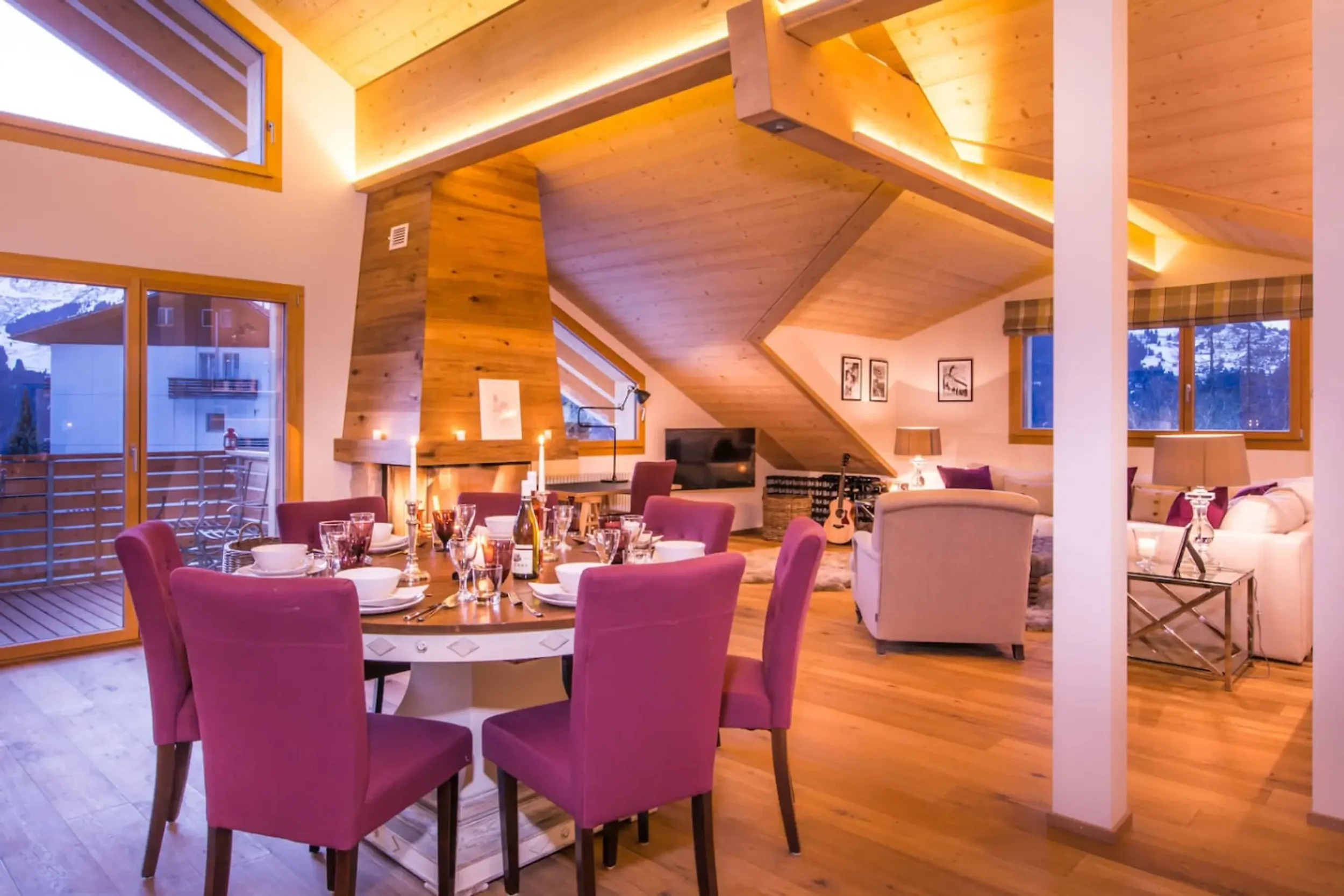 A cozy dining area with a set table, pink chairs, a fireplace, wooden ceiling and walls, large windows, and a scenic mountain view outside.
