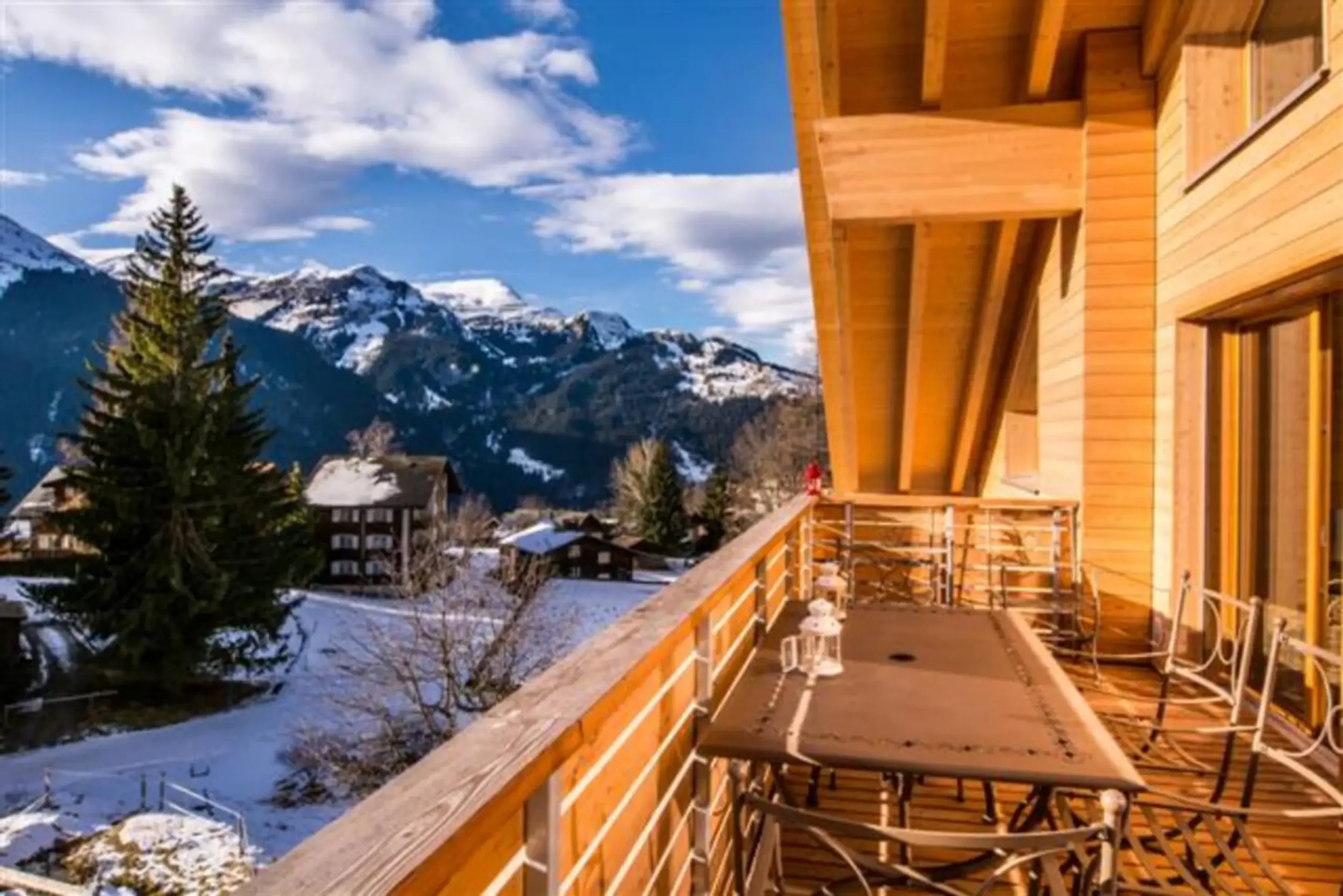 View from a balcony of a wooden chalet overlooking snow-covered mountains and neighboring chalets in a winter scene.