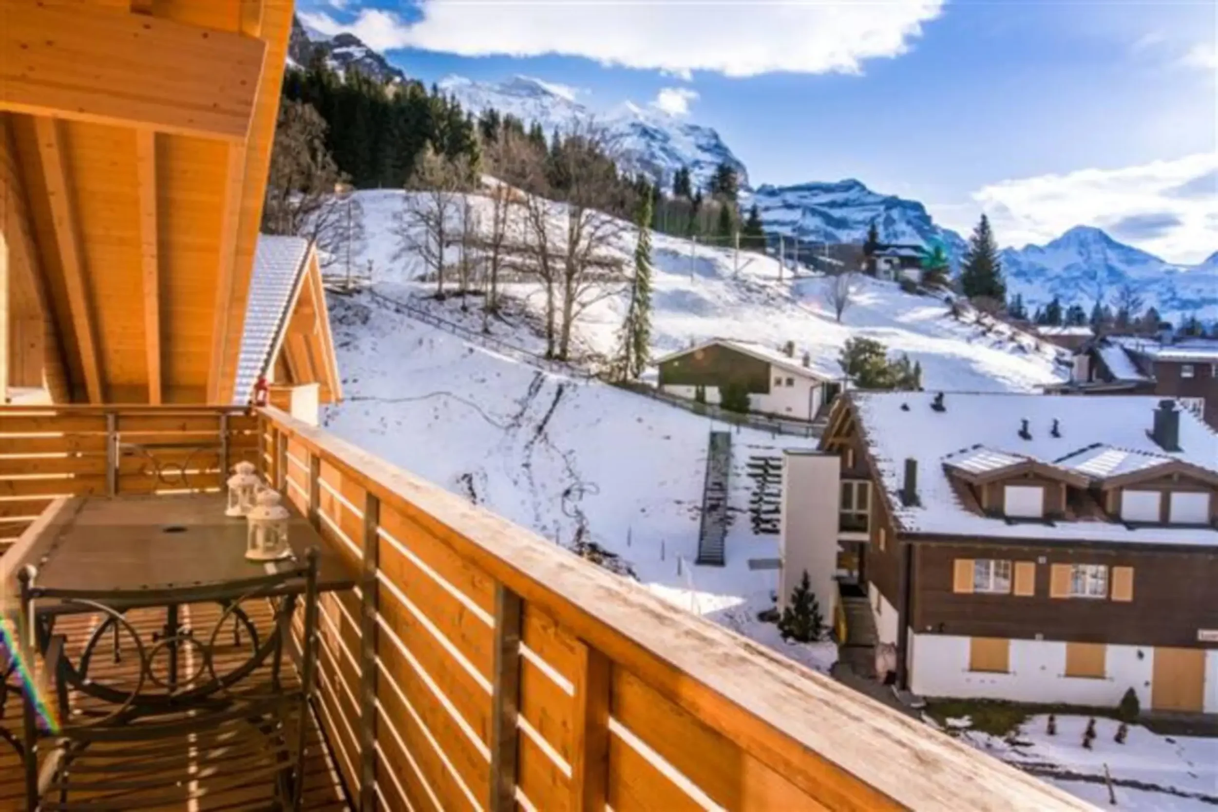 View from a wooden balcony overlooking snow-covered houses, trees, and mountain peaks under a blue sky with clouds.