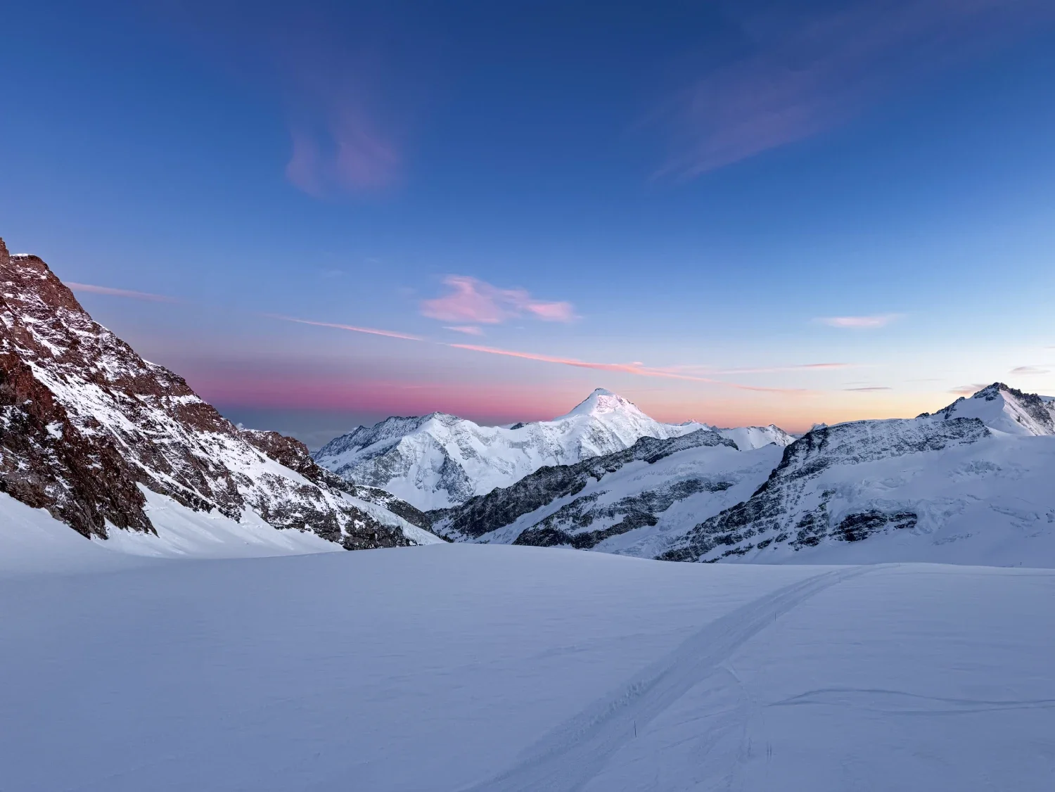 Snow-covered mountains under a colorful sky at dusk or dawn with tracks in the snow in the foreground.