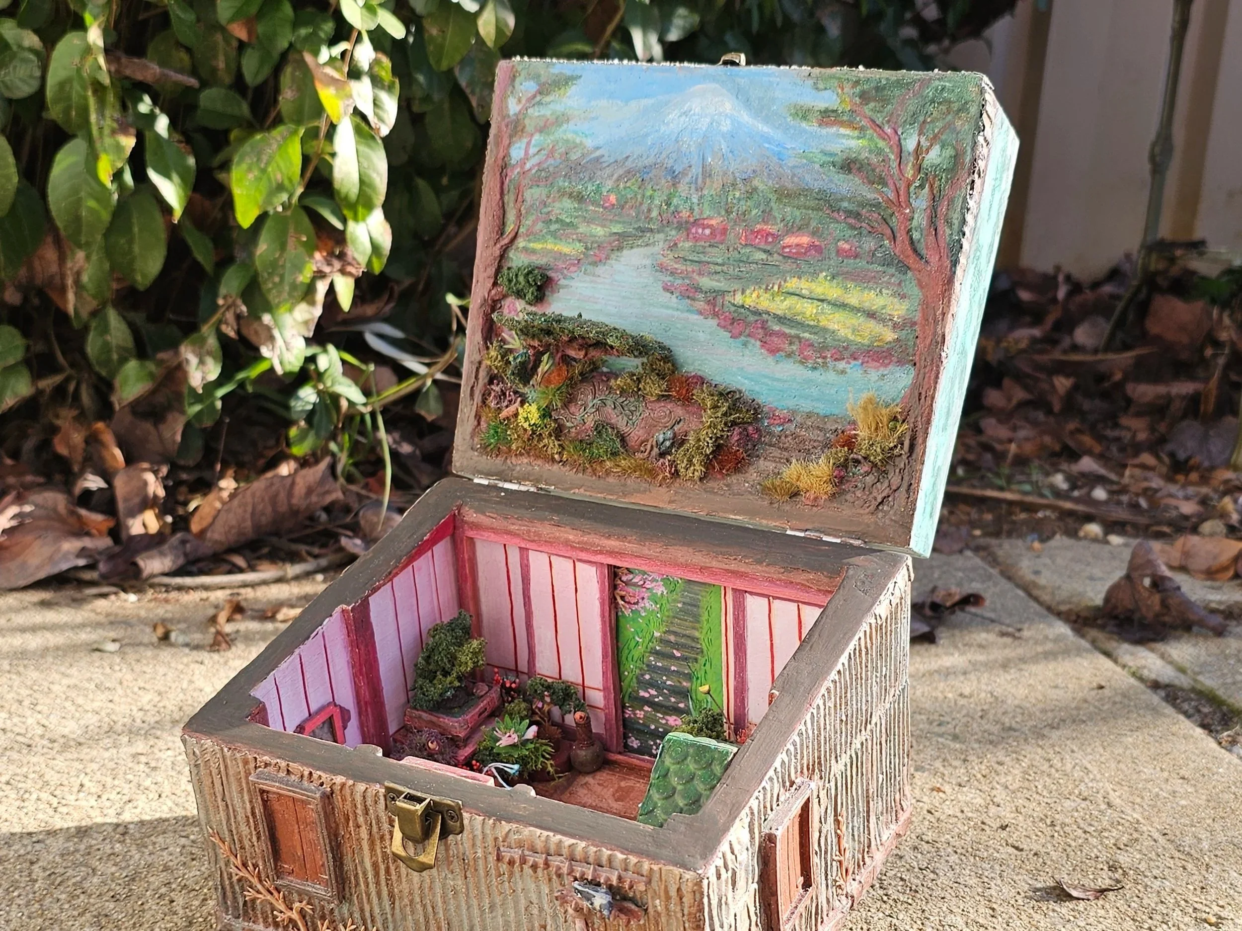 A small, decorative wooden house with painted walls and miniature plants inside. The house has a pink and green color scheme, with the roof and interior walls painted in bright colors. It is placed outdoors on a surface with foliage in the background.