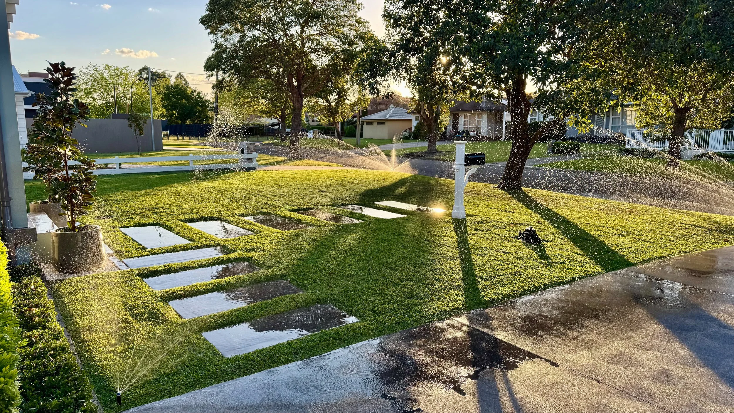 An automated sprinkler system watering a vibrant, neatly manicured lawn with stepping stones in a well-maintained residential front yard.