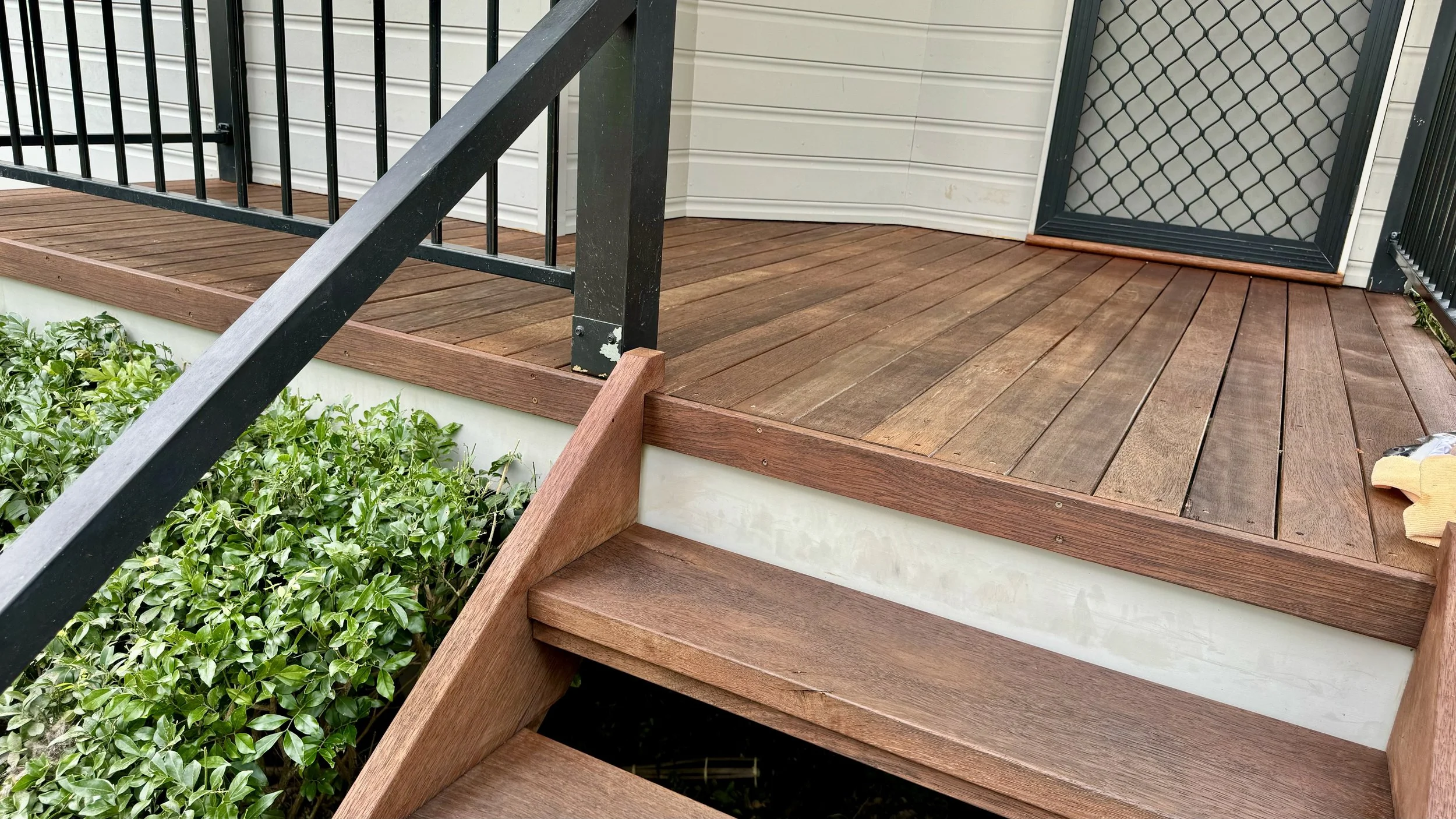 Close-up view of a freshly sanded, treated, and stained timber deck entrance staircase with black metal handrails.