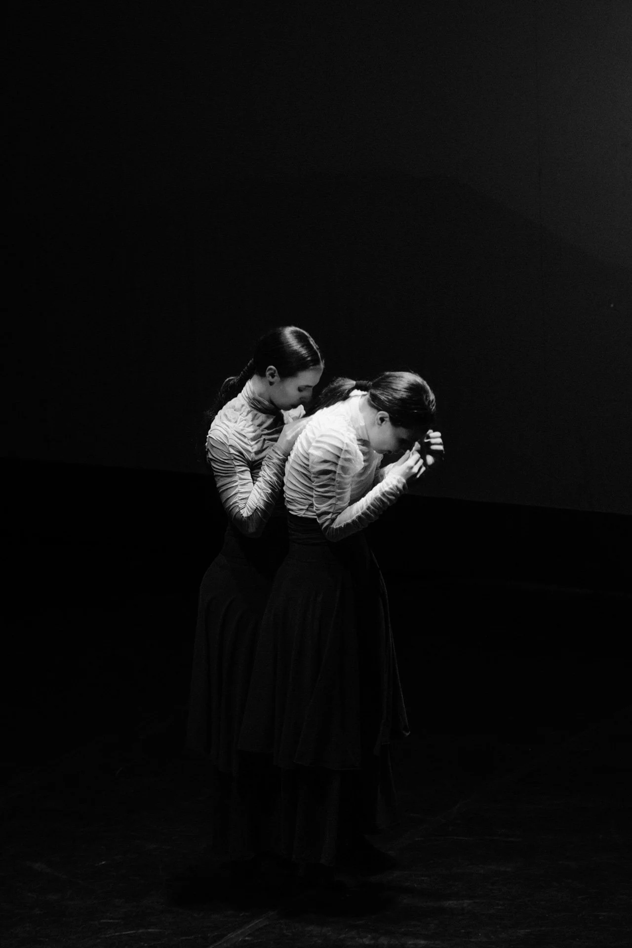 Two women dressed in striped tops and long skirts are performing a dance on stage with a dark background, their heads bowed and hands clasped in front of their faces.