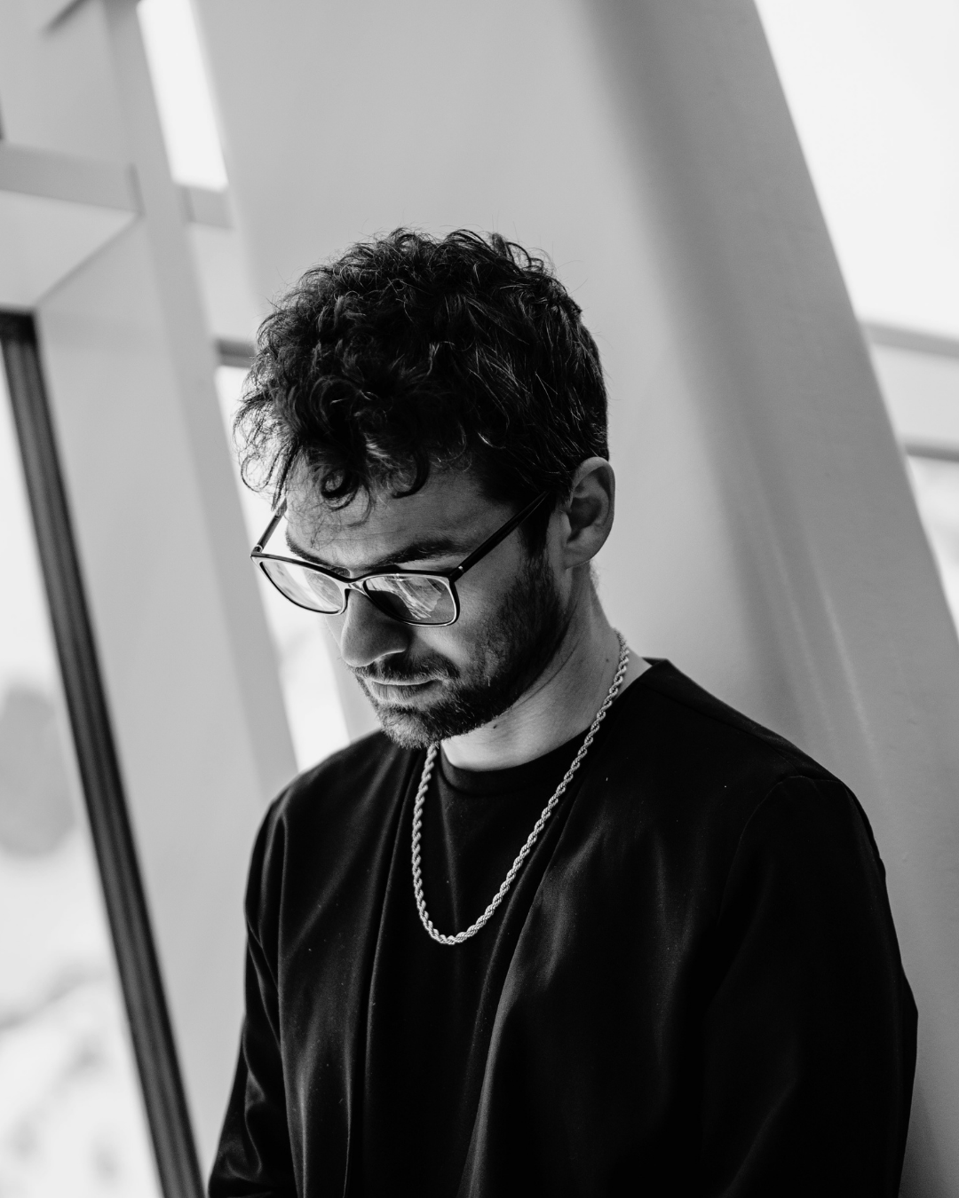 A black-and-white photo of a man with dark curly hair, glasses, and a beard, looking down thoughtfully, wearing a black shirt and a chain necklace, in an indoor setting near a window.