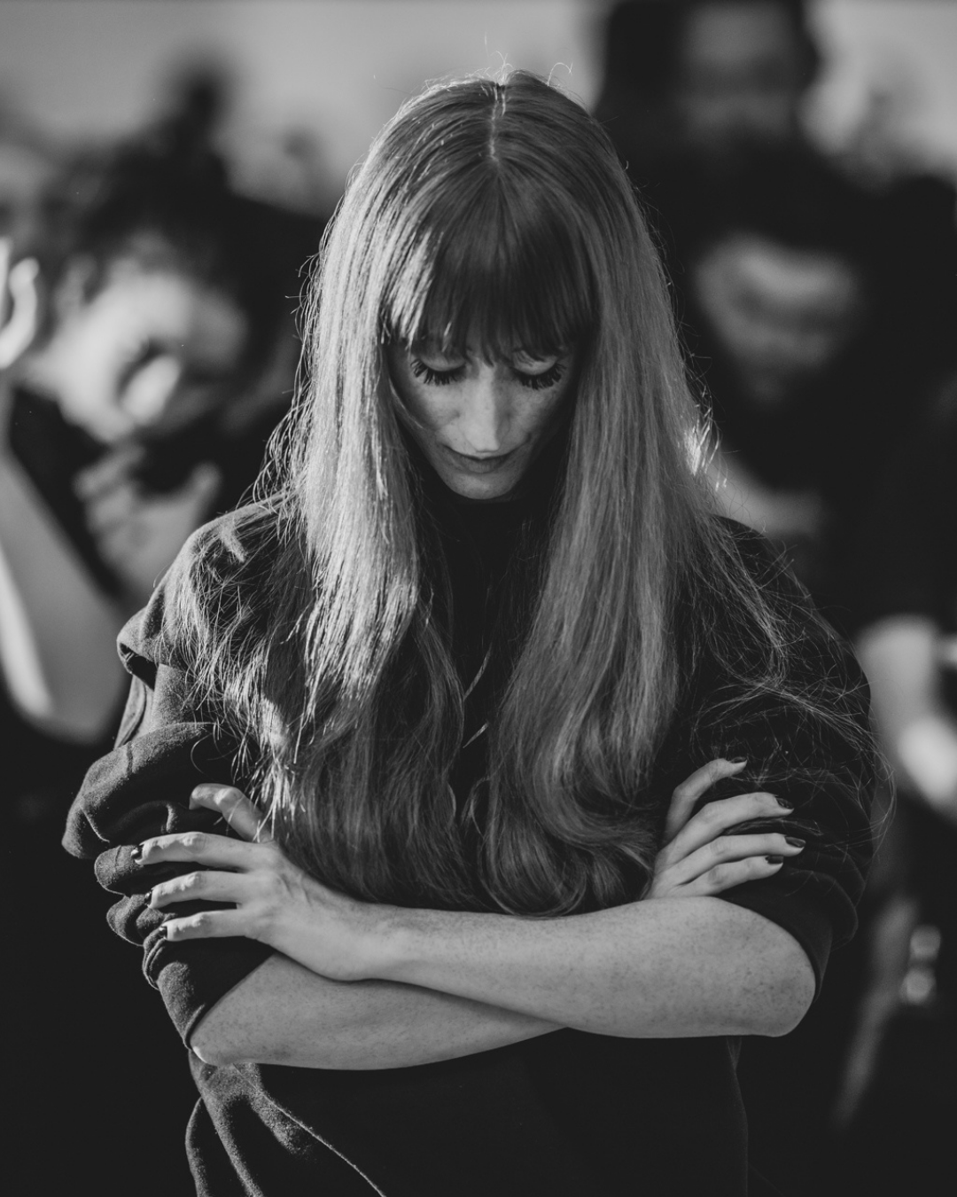 A black and white photo of a woman with long hair and bangs, looking down with her arms crossed. Blurred people are in the background.