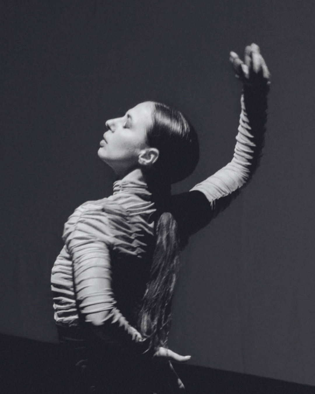 Black and white photo of a young woman with closed eyes and her head tilted back, raising one arm with a closed fist, wearing a striped long-sleeve top and the other hand on her hip, against a dark background.