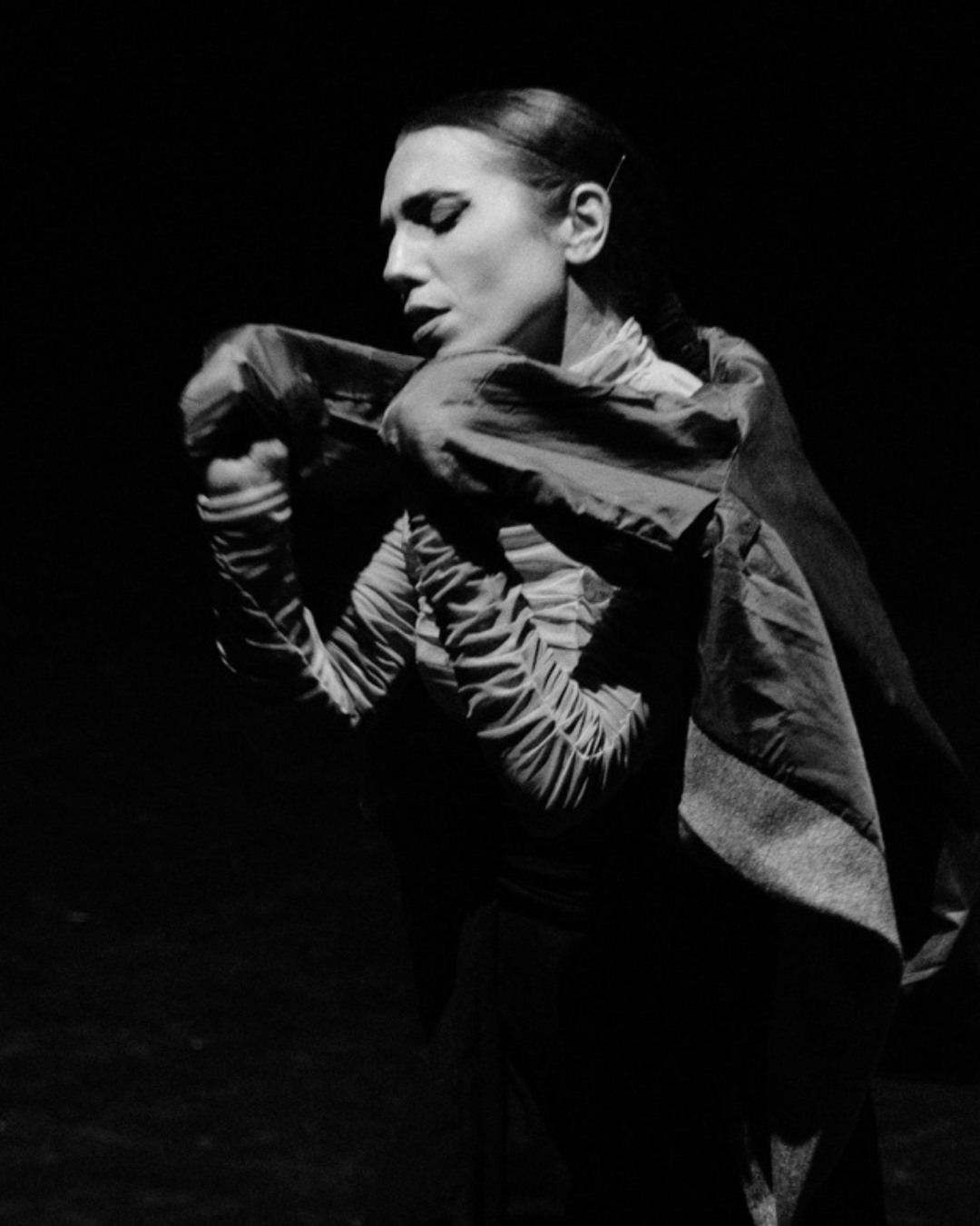 A black and white photo of a woman with slicked-back hair, wearing a jacket with striped sleeves, posing with her eyes closed and arms crossed in front of her face against a dark background.