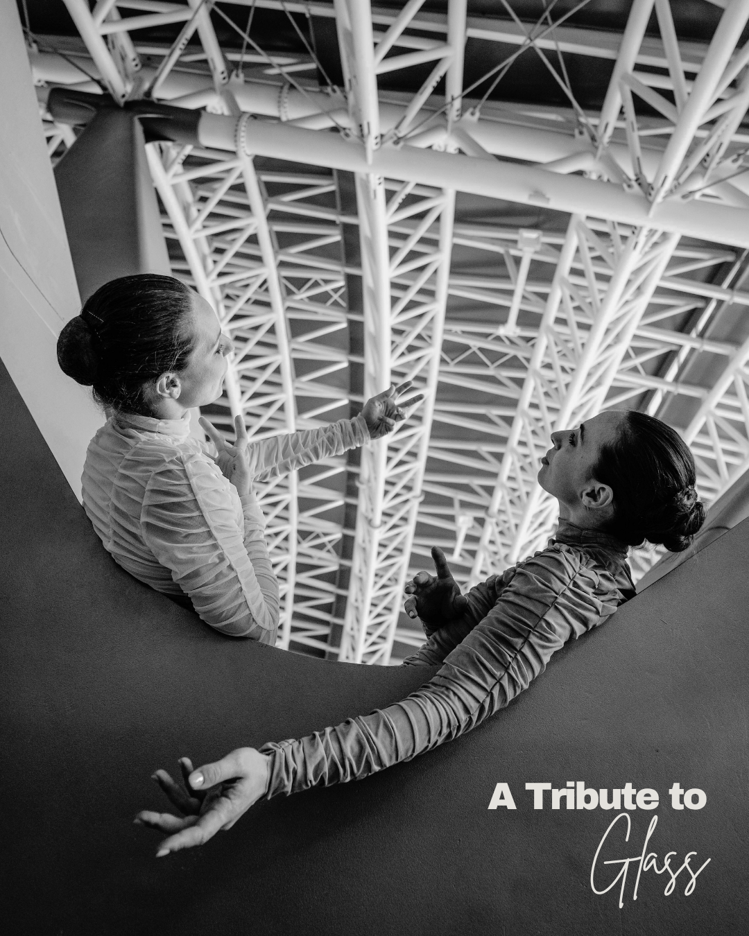 Two women in dance or gymnastic attire sitting on a curved surface, engaging in a conversation in a modern building with a complex network of metal beams above them. The image is in black and white, and the text 'A Tribute to Glass' is written at the bottom right corner.