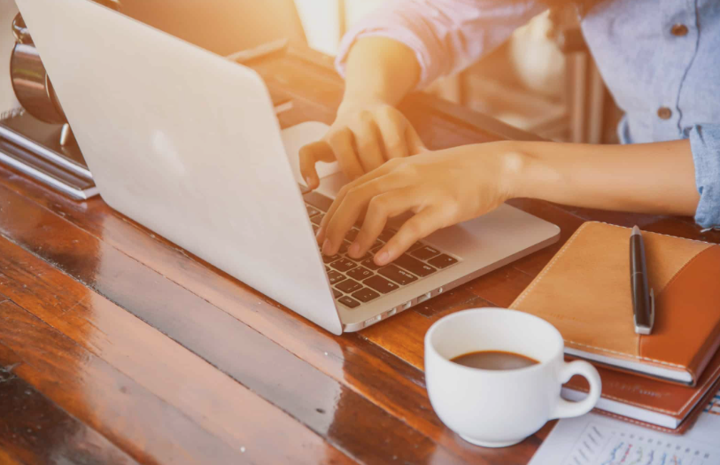 Person typing on a silver laptop at a wooden desk, with a white coffee mug, a notebook, and a pen nearby.