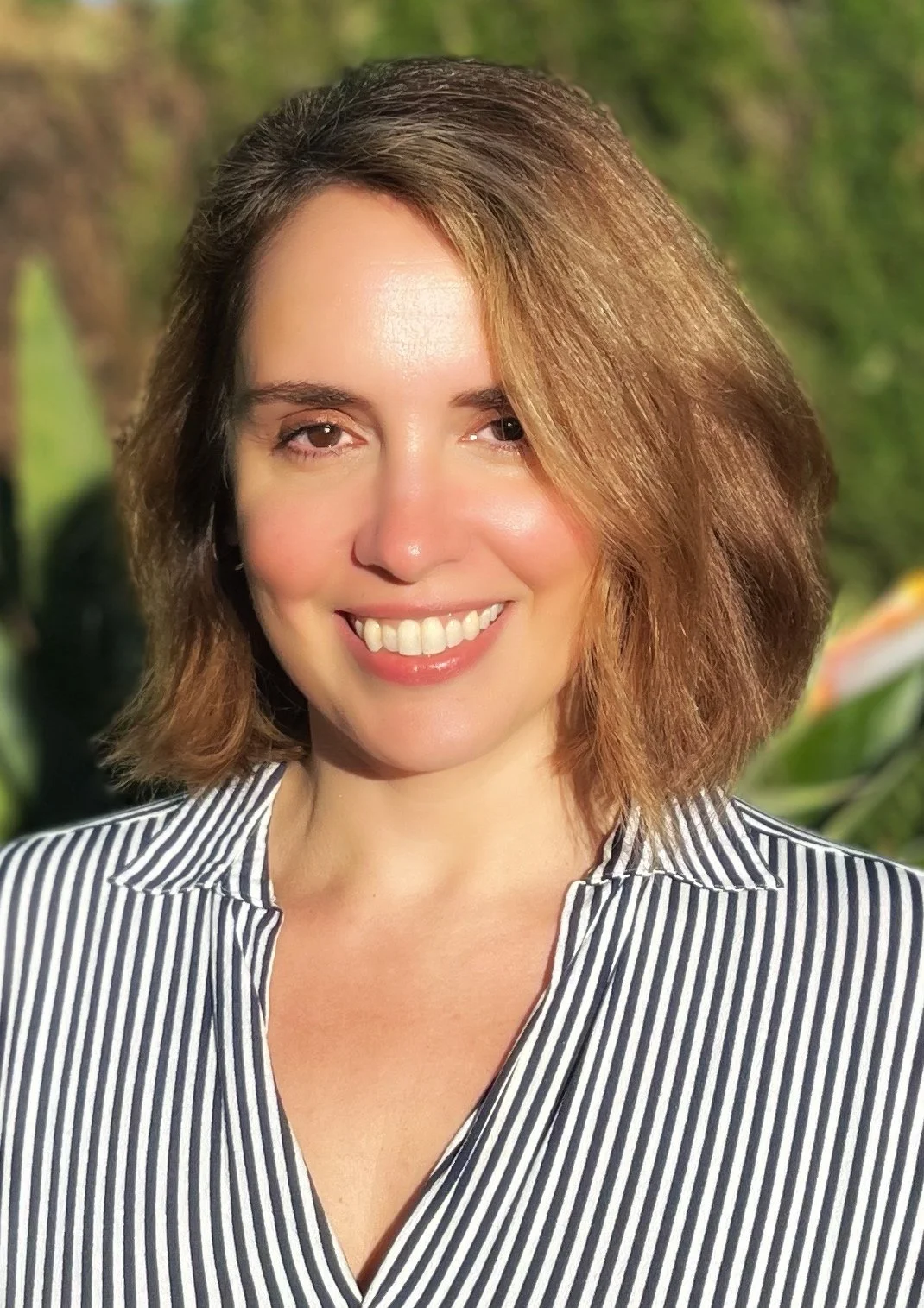 A smiling woman with shoulder-length light brown hair, wearing a black and white striped shirt, outdoors with a blurred green background.