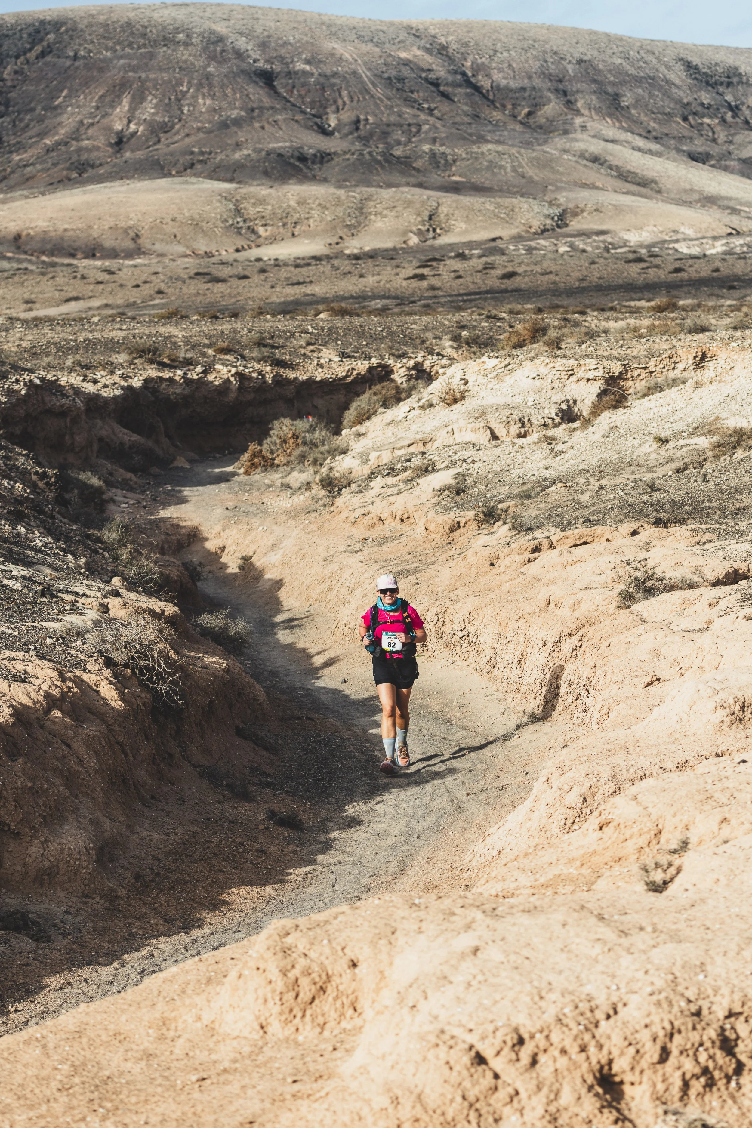 A person wearing athletic gear, including a pink backpack and a white cap, running along a narrow dirt trail in a desert landscape with rocky hills in the background.