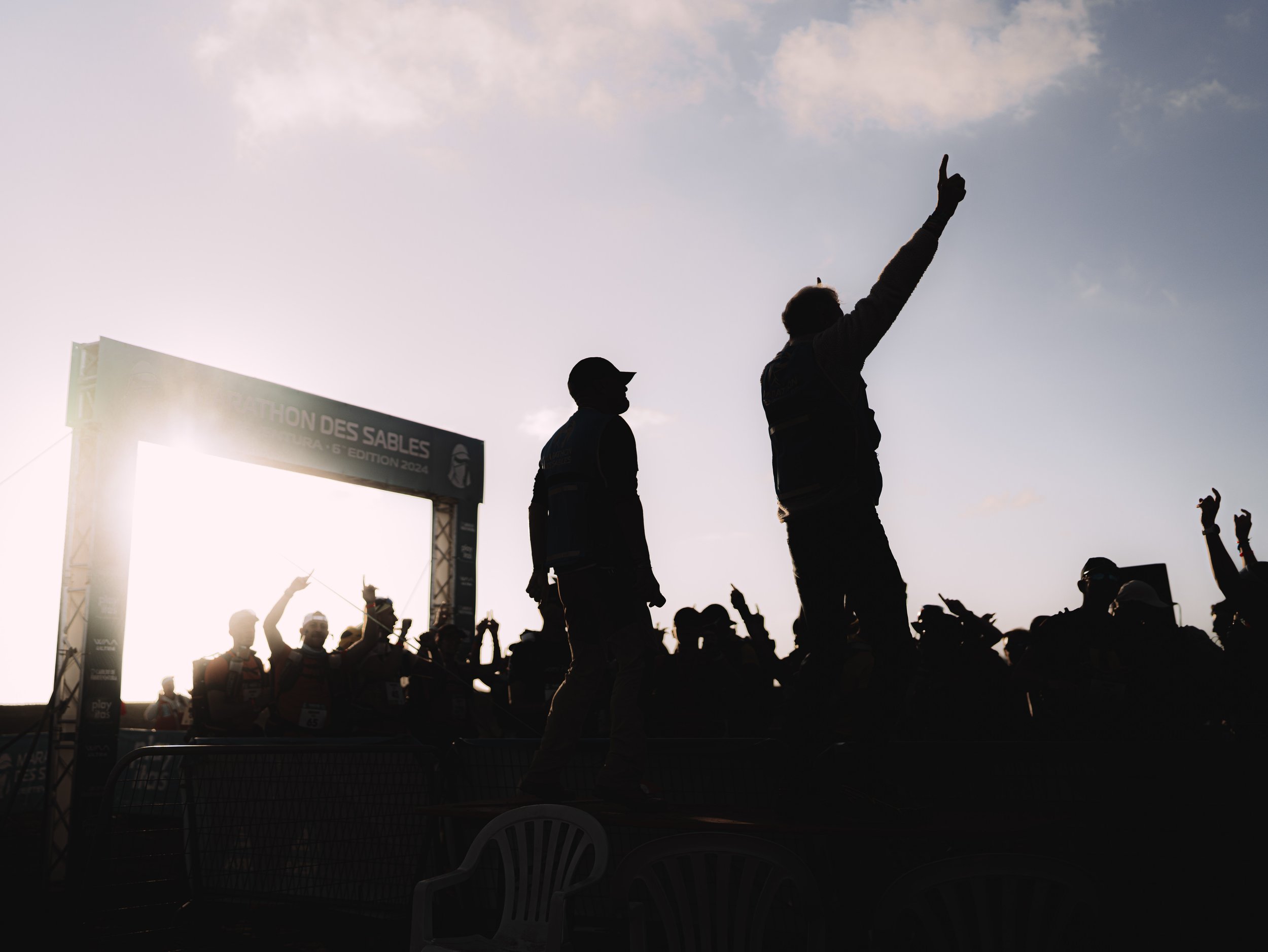 Silhouettes of people, some raising their hands and pointing, gather at the finish line of a marathon race during sunset, with a large arch reading 'MARATHON DES SABLES 2024' in the background.