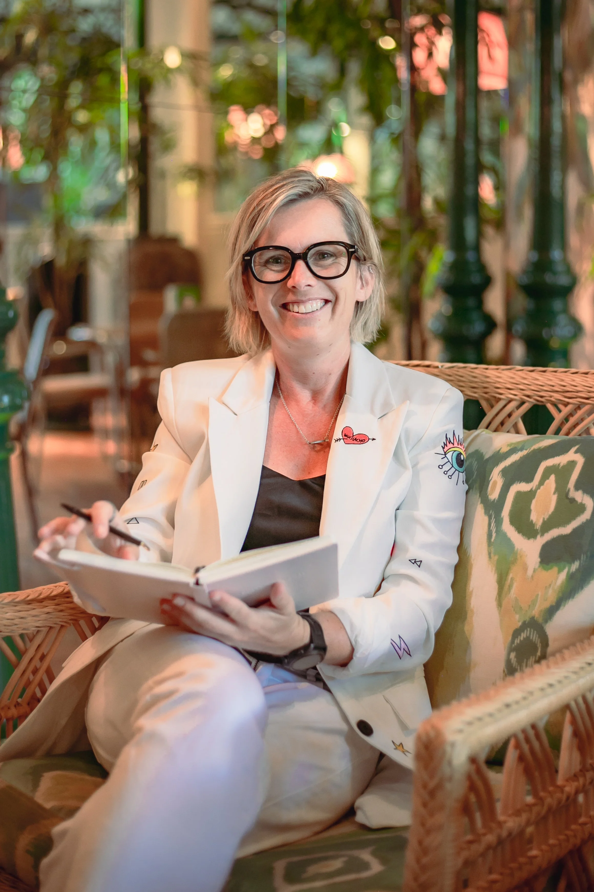 A cheerful woman with blonde hair and glasses, wearing a white blazer with colorful patches, sitting on a wicker chair with a notebook in her lap in an outdoor setting with greenery and warm lighting in the background.