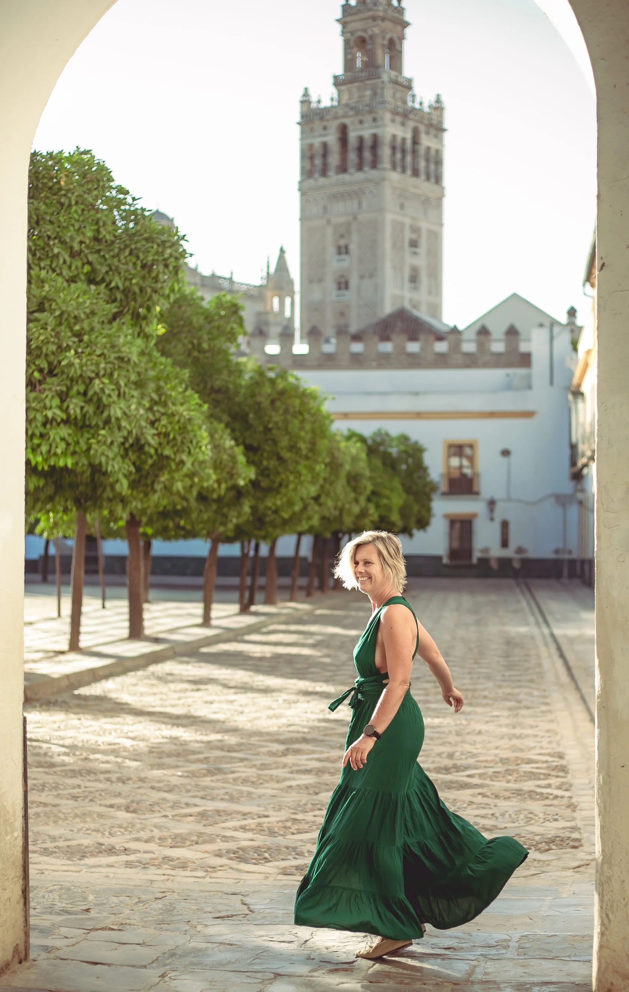 A woman in a flowing green dress walking through an arched stone doorway with a castle tower in the background during daytime.