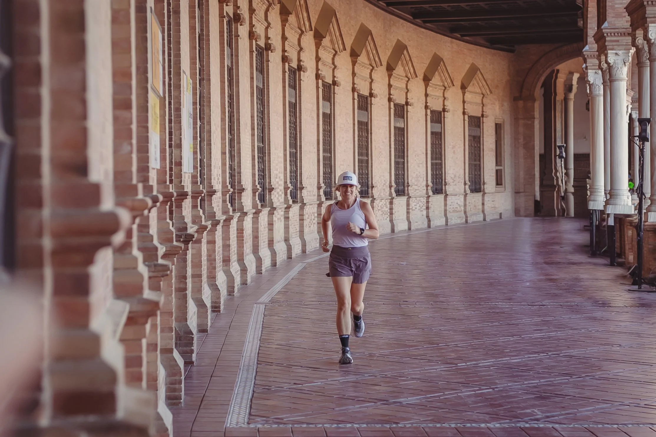 A woman jogging along a covered brick walkway with ornate columns and large windows on a sunny day.