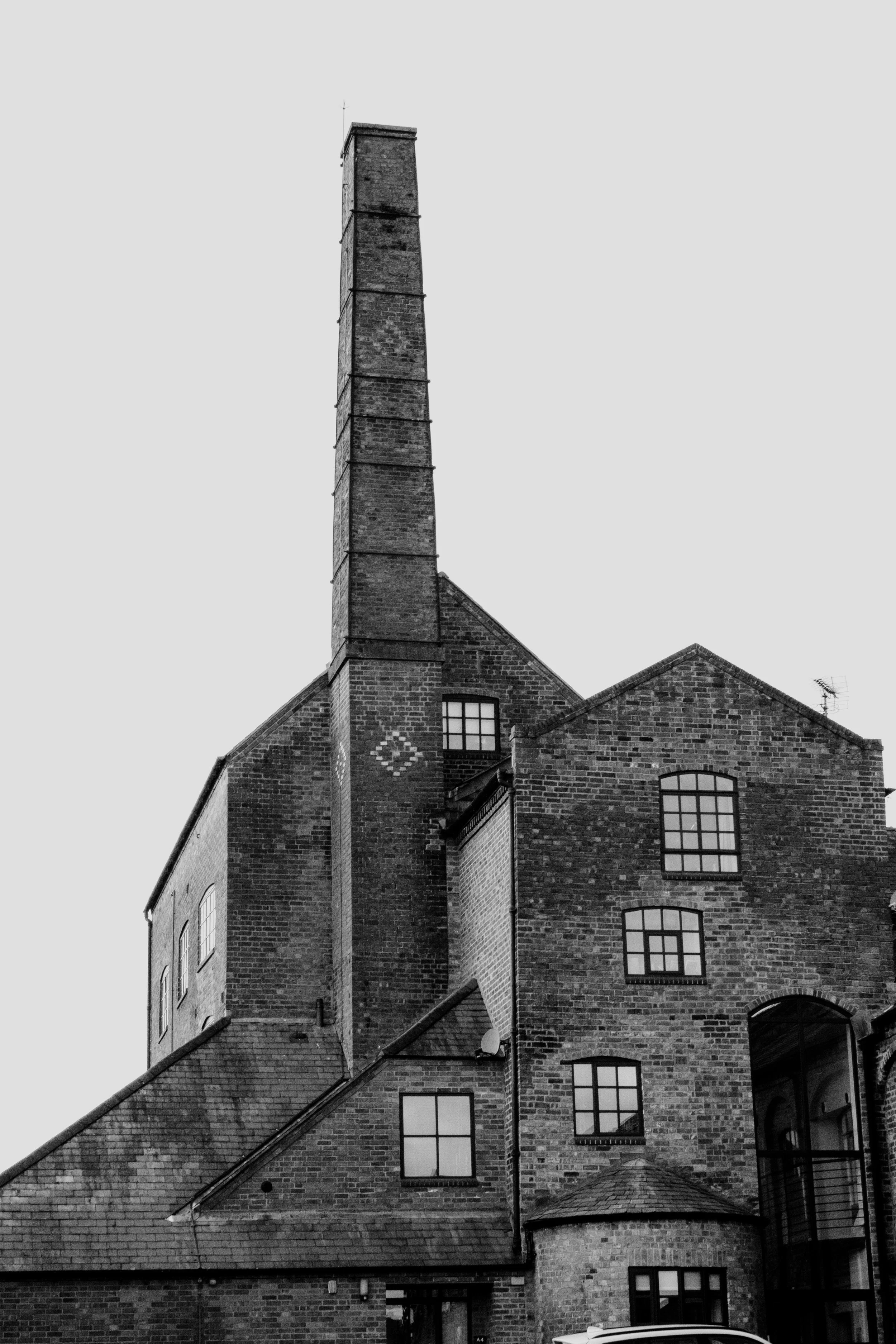 A tall brick building with multiple windows and a large chimney or smokestack extending upward, against a plain sky.