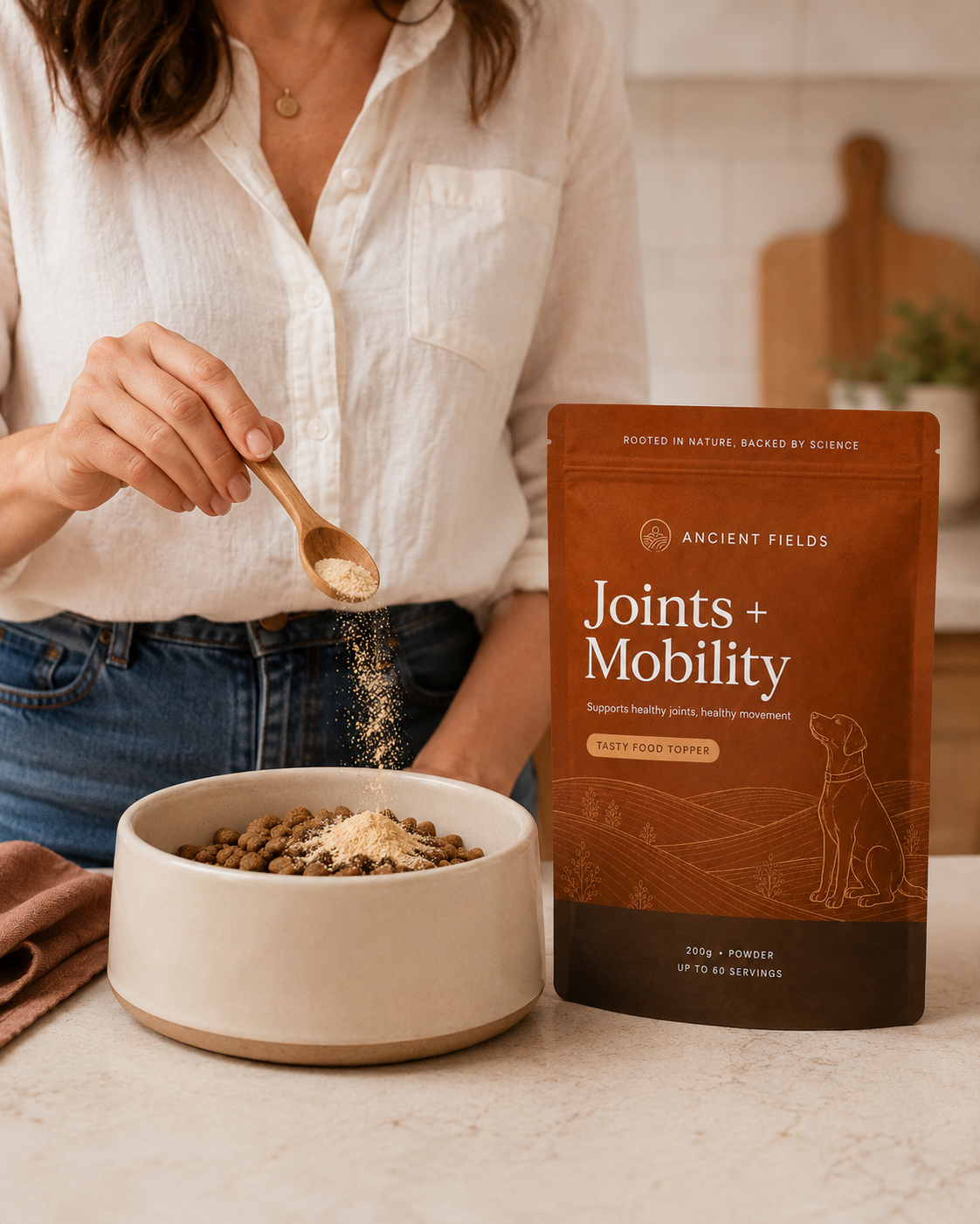 A woman is sprinkling powder from a wooden spoon onto dry dog food in a ceramic bowl. Next to her, there is a package of Ancient Fields Joints + Mobility supplement for dogs.