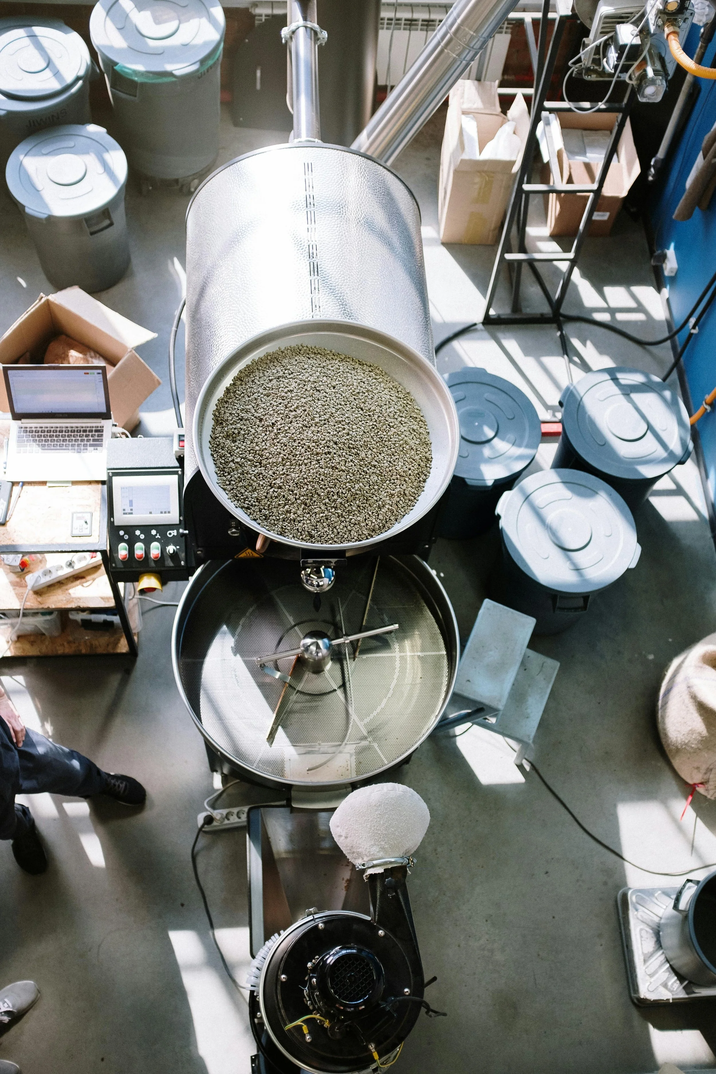Overhead view of a laboratory setup with a metal grinding or milling machine, a laptop, various control panels, and trash bins, all illuminated by natural sunlight.
