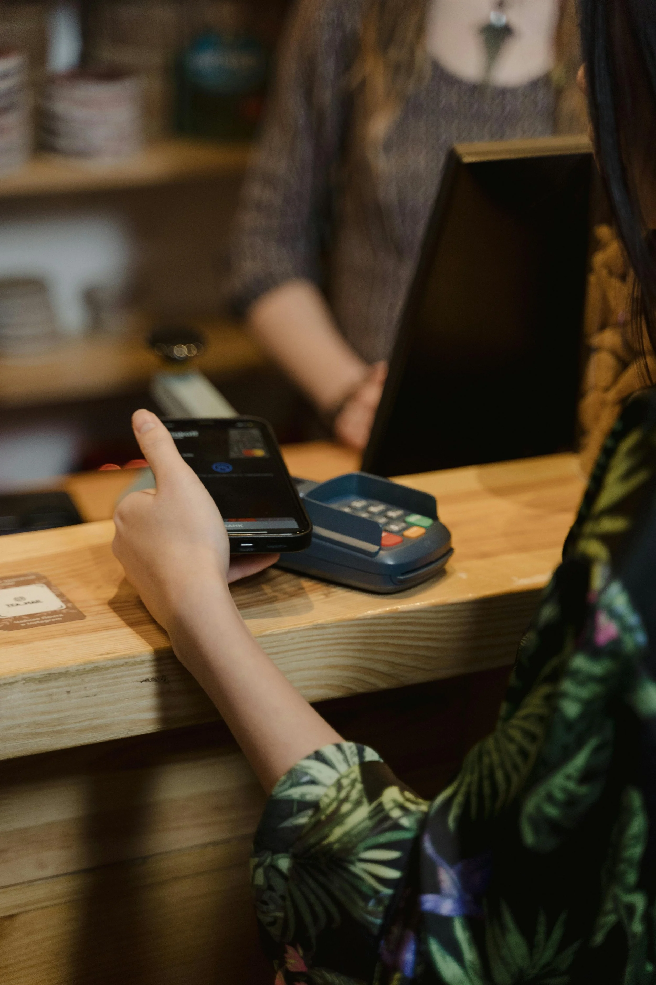 A person with floral patterned clothing using a smartphone at a wooden counter, with a card reader device, in what appears to be a café or restaurant. There is a person with a gray sweater in the background, and shelves with dishes.