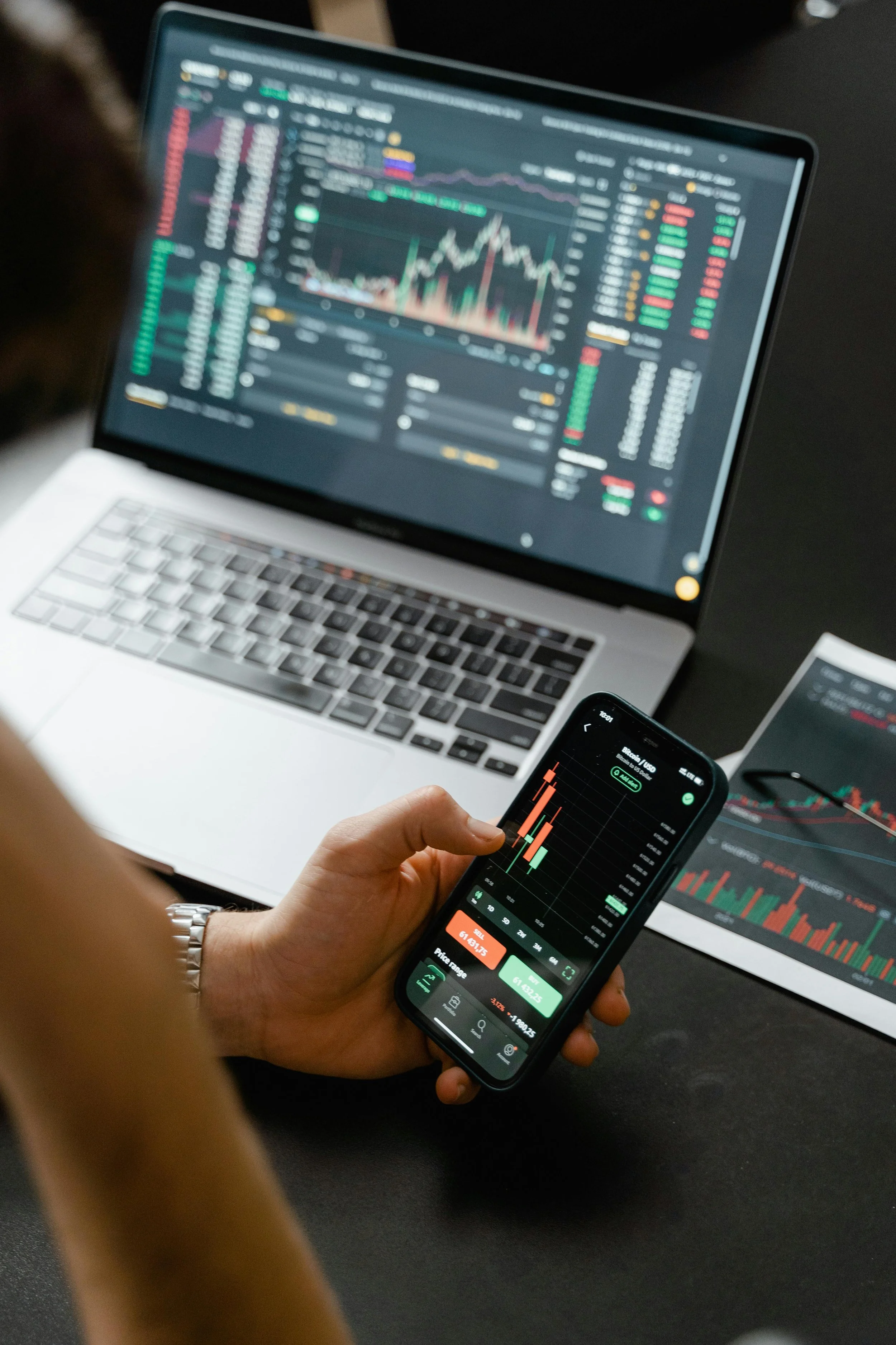 Person holding a smartphone displaying a stock trading app, with a laptop on a black table showing financial charts and data.