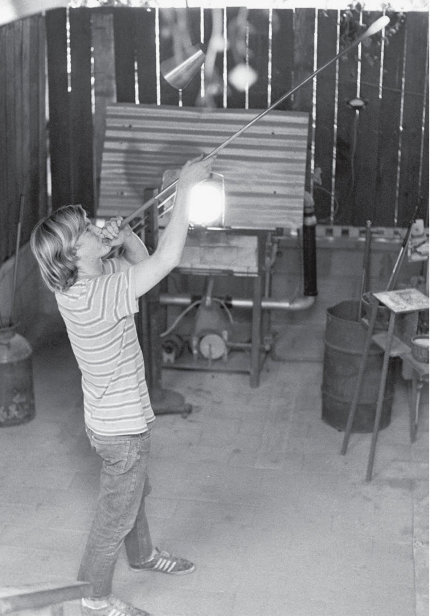 Black-and-white photo of a person shaping molten glass with a blowpipe inside a workshop.