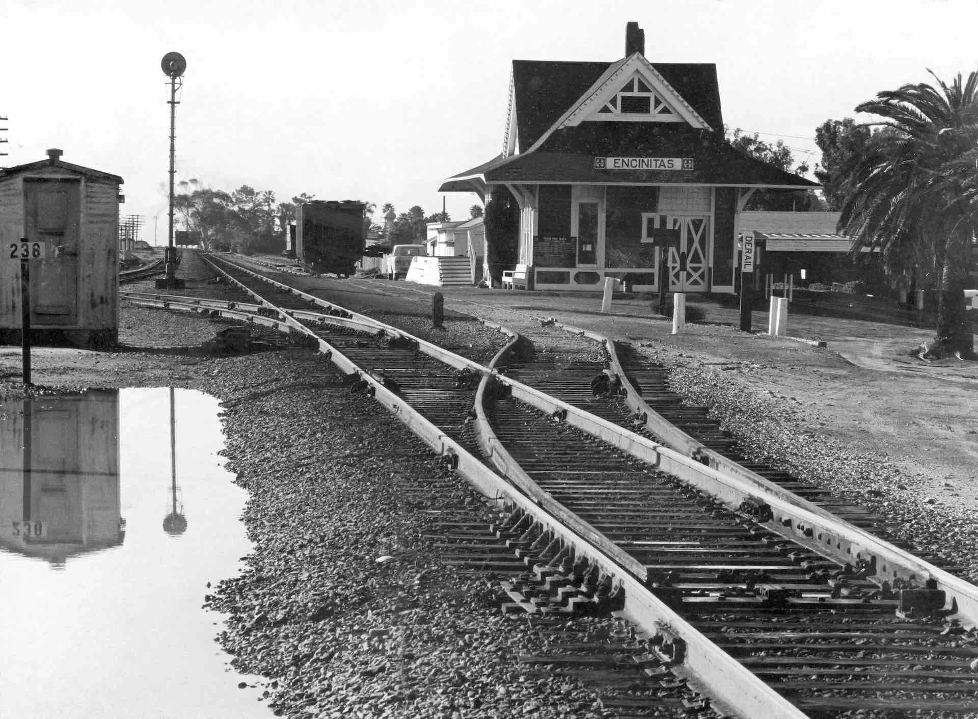 Black-and-white photo of the historic Encinitas train station beside railroad tracks and a large palm tree.
