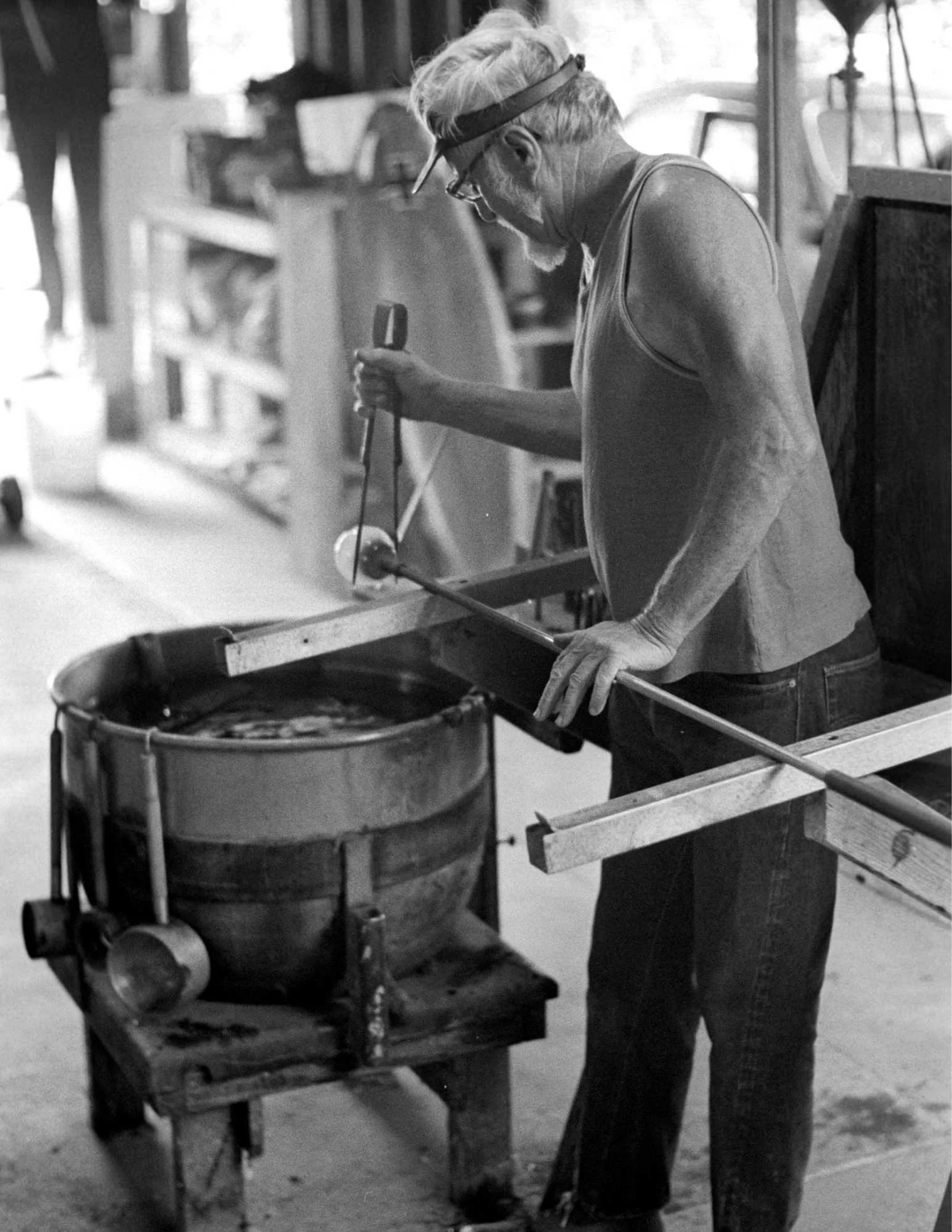 Black-and-white photo of an older artisan shaping molten glass in a workshop.